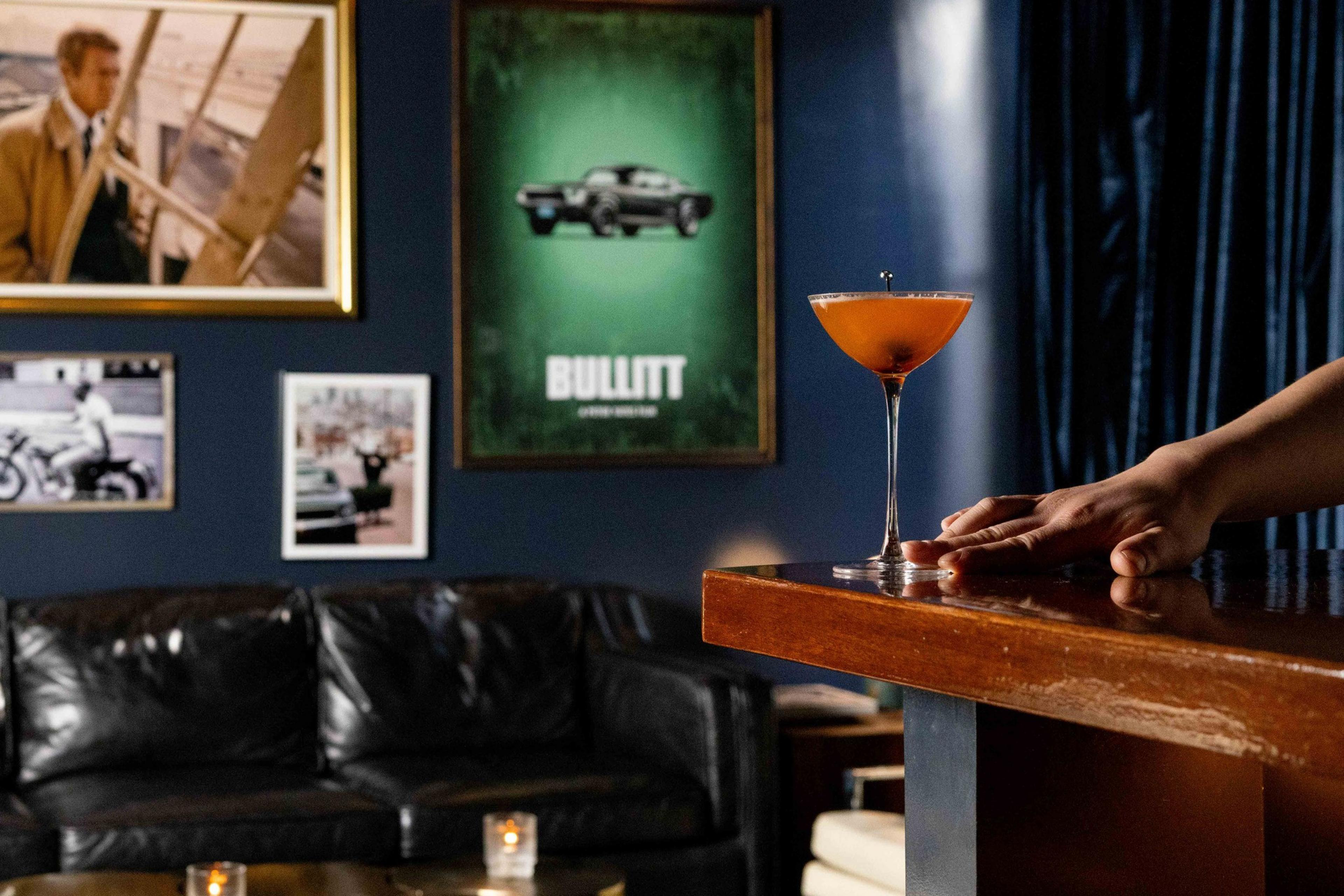 A cozy room with a black leather couch, framed movie posters, and photos on dark blue walls. A hand rests near a martini glass with an orange drink on a wooden table.