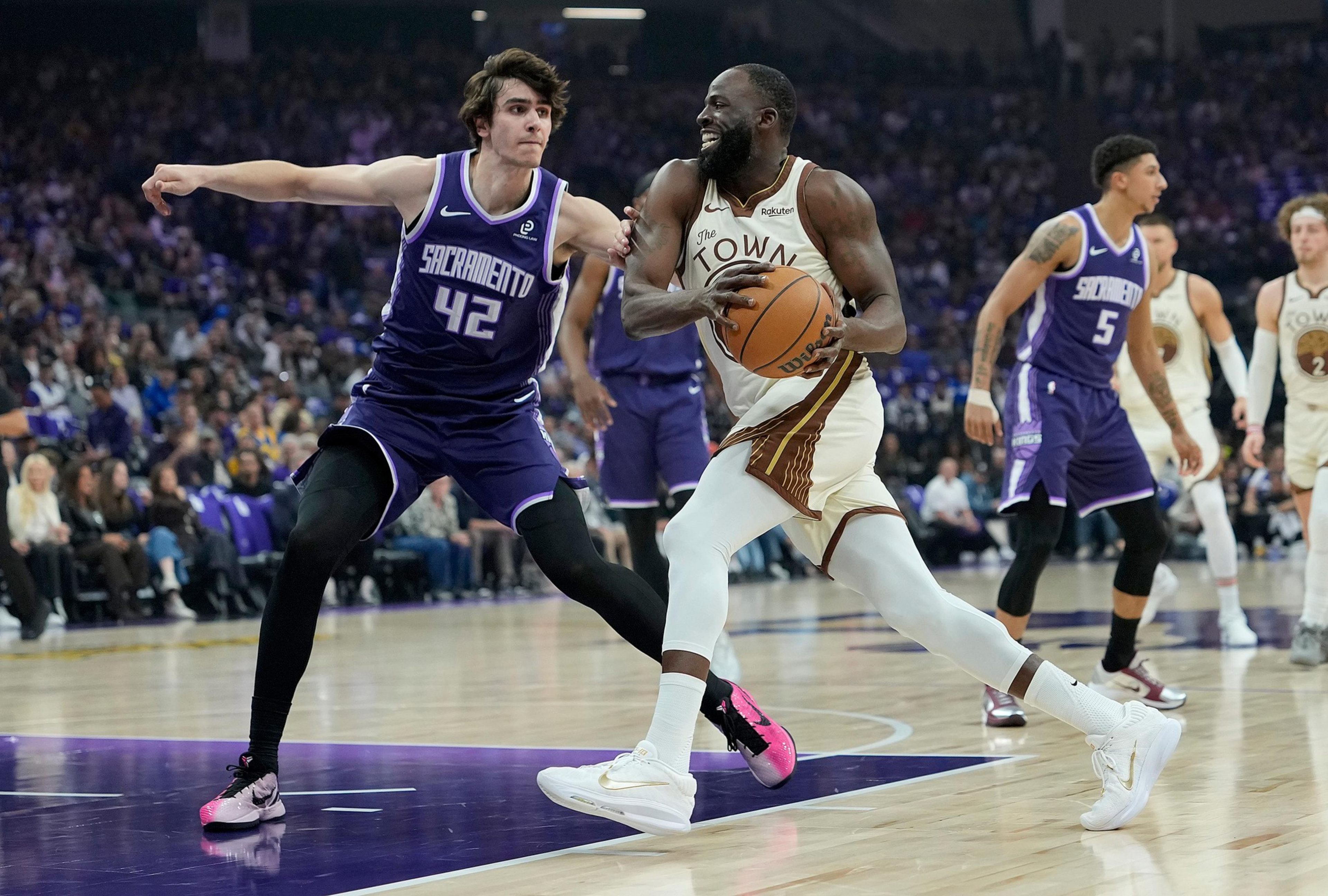 A basketball player in a white uniform dribbles while being closely guarded by a defender in a purple Sacramento Kings uniform during a game.