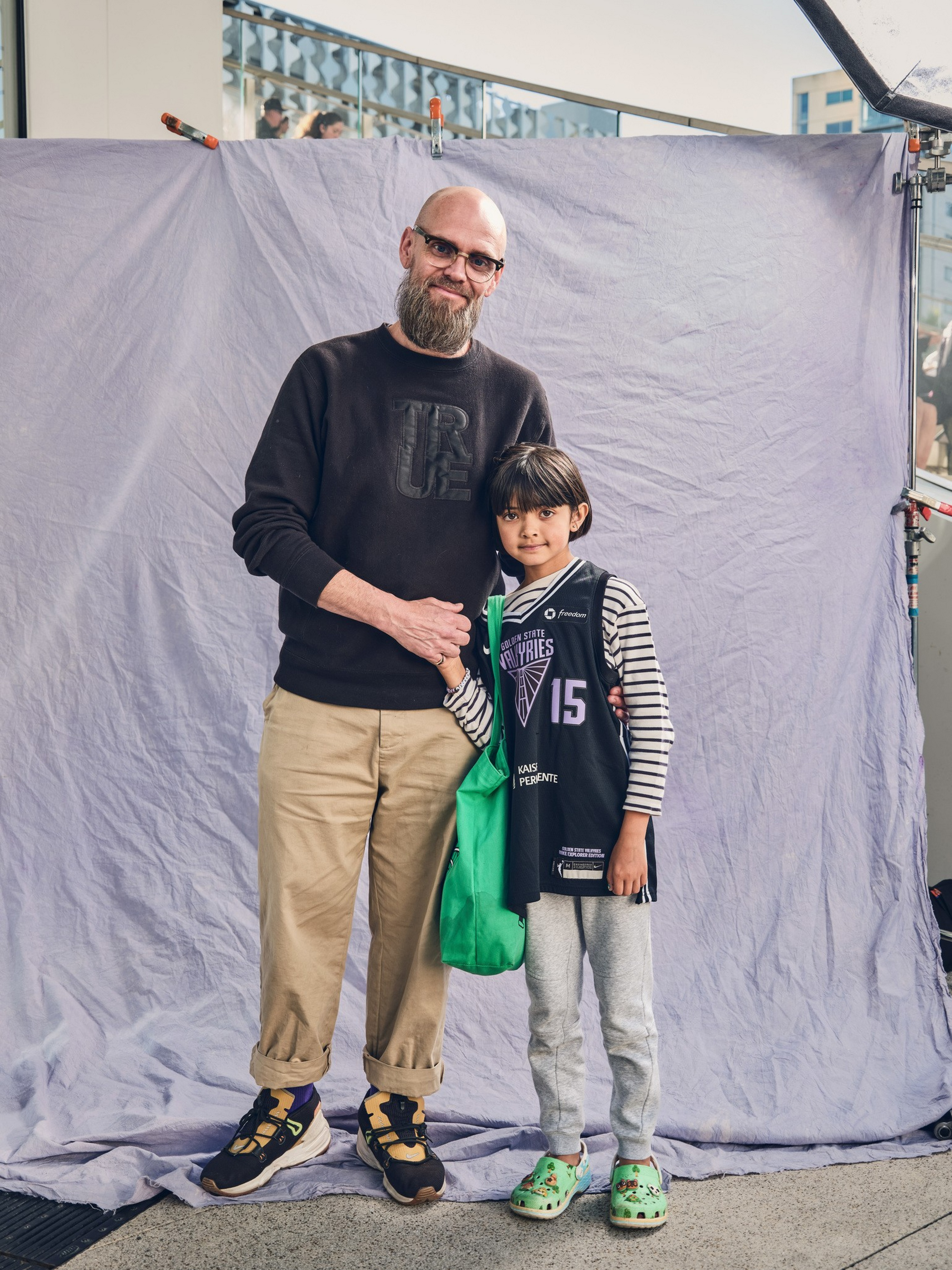 A bearded man in glasses and beige pants stands beside a child in a basketball jersey and gray sweatpants, both holding hands and smiling.