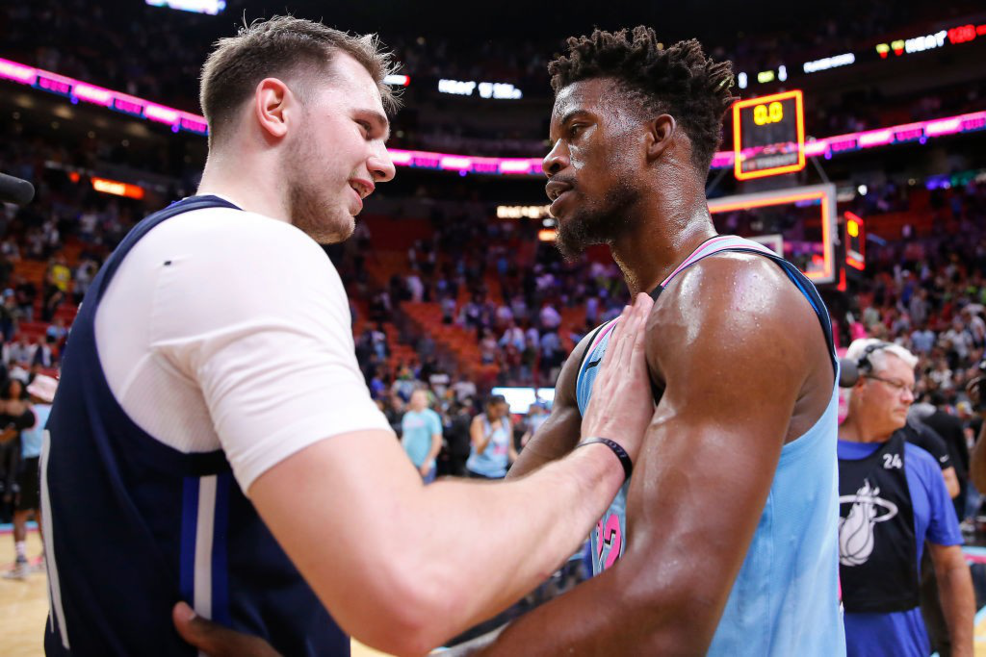 Two basketball players, one in a white and blue uniform and the other in light blue and pink, are sharing a moment on the court, surrounded by a vibrant crowd.