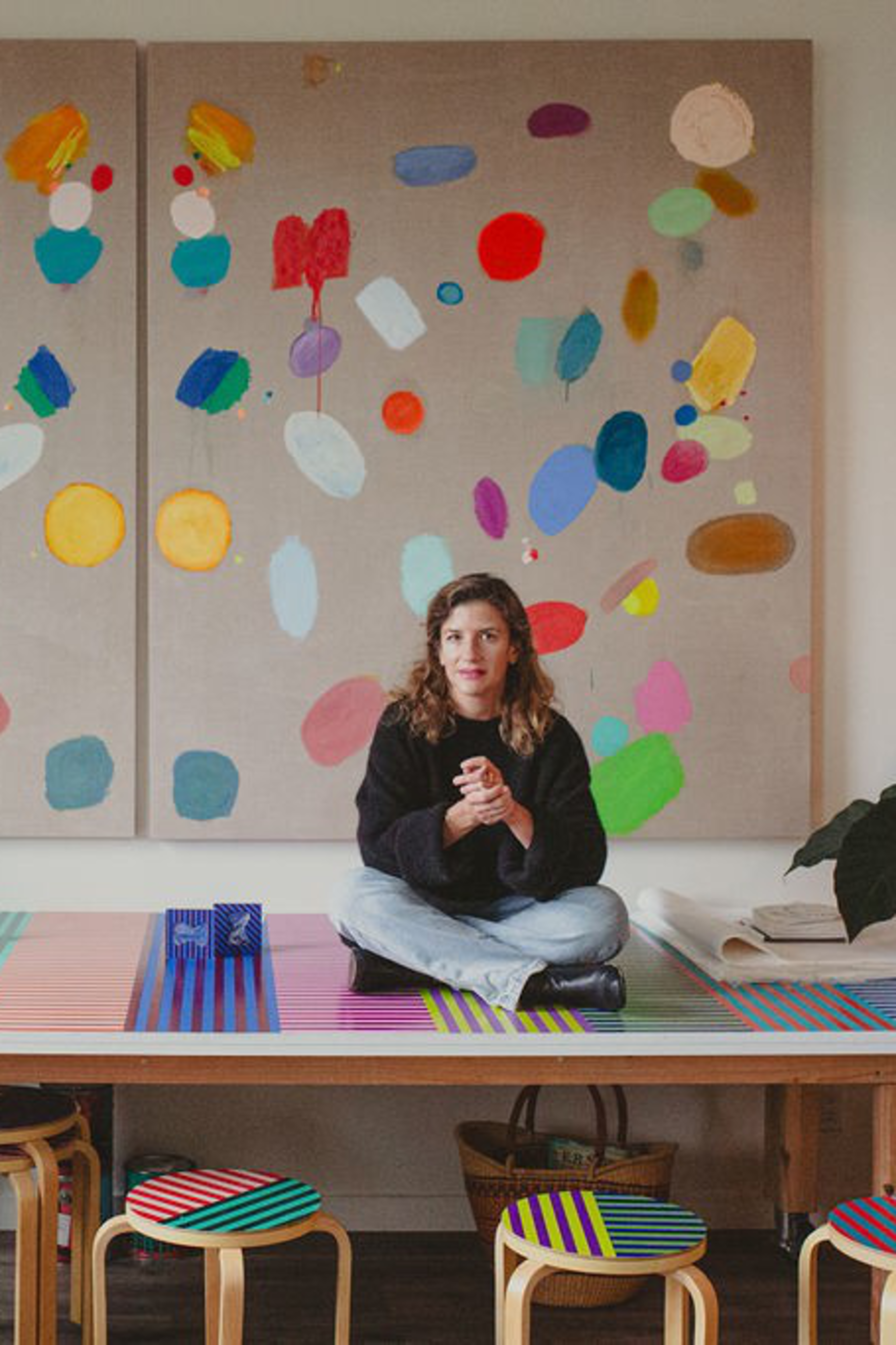 A person sits cross-legged on a colorful table with striped stools, framed by vibrant abstract art pieces and surrounded by lush potted plants.