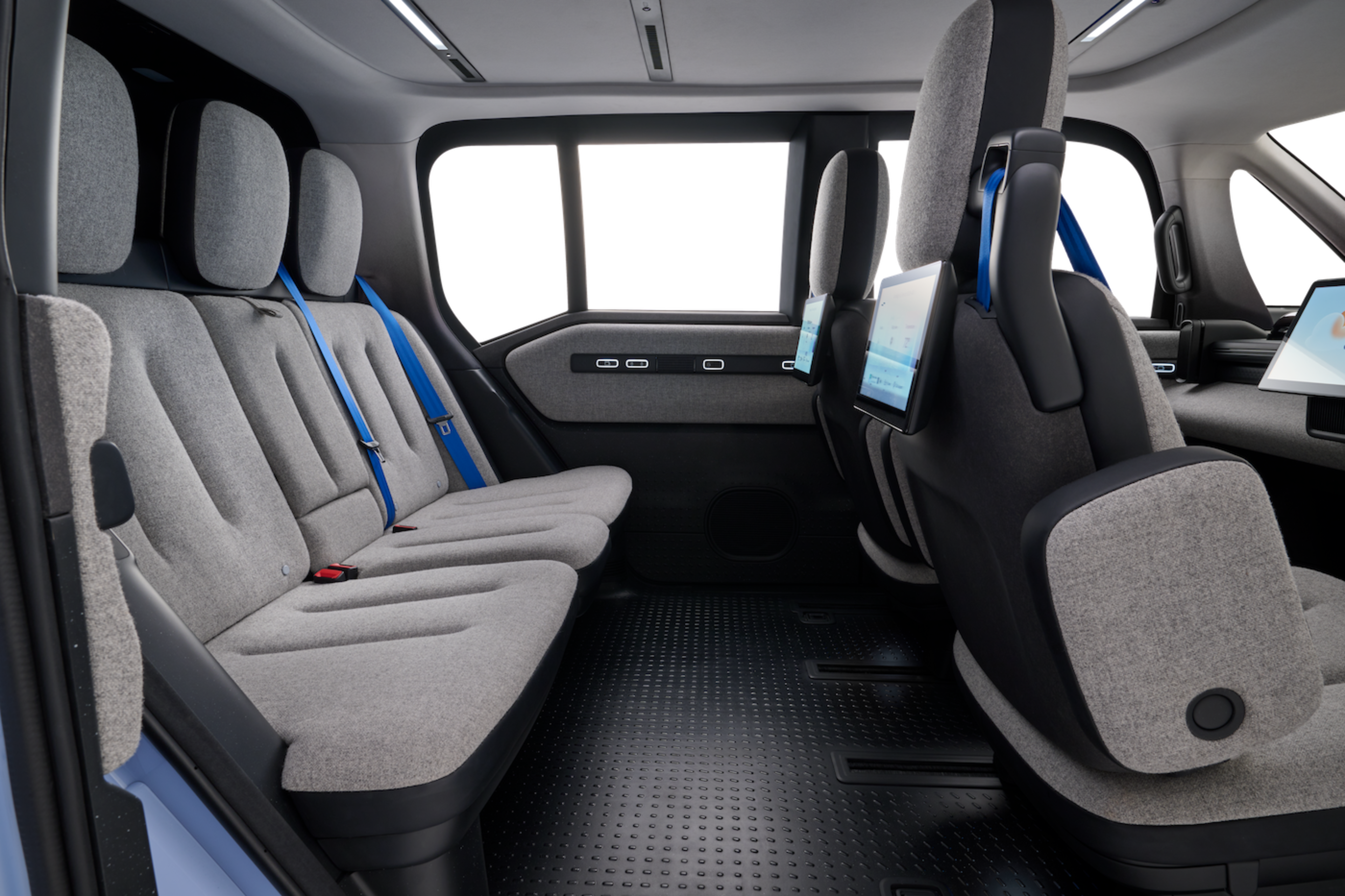 A car interior with gray fabric seats, blue seat belts, and digital screens mounted on the back of the front seats, plus large windows.
