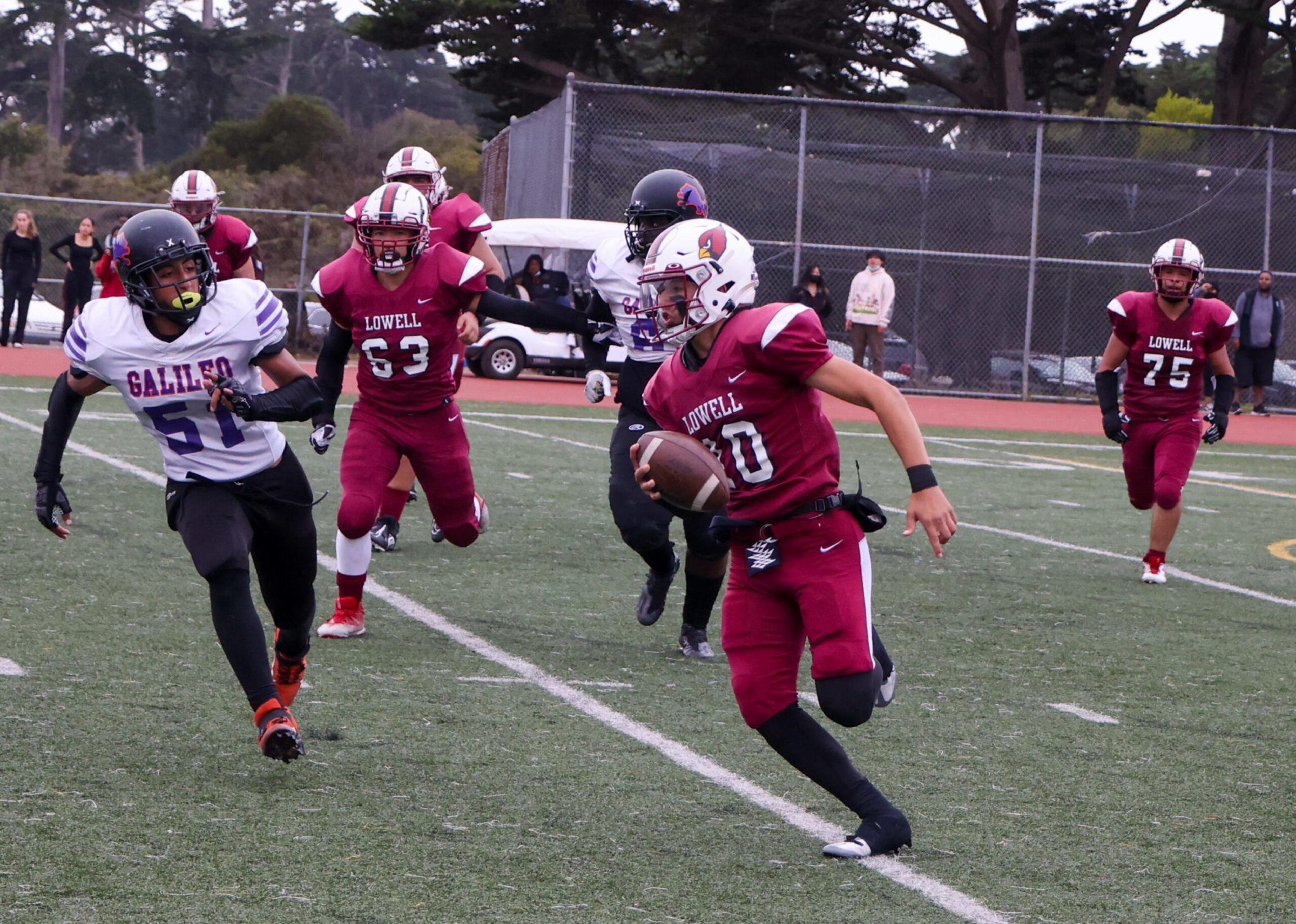A football player in a maroon Lowell uniform runs with the ball while players in white and maroon chase him on a green field.