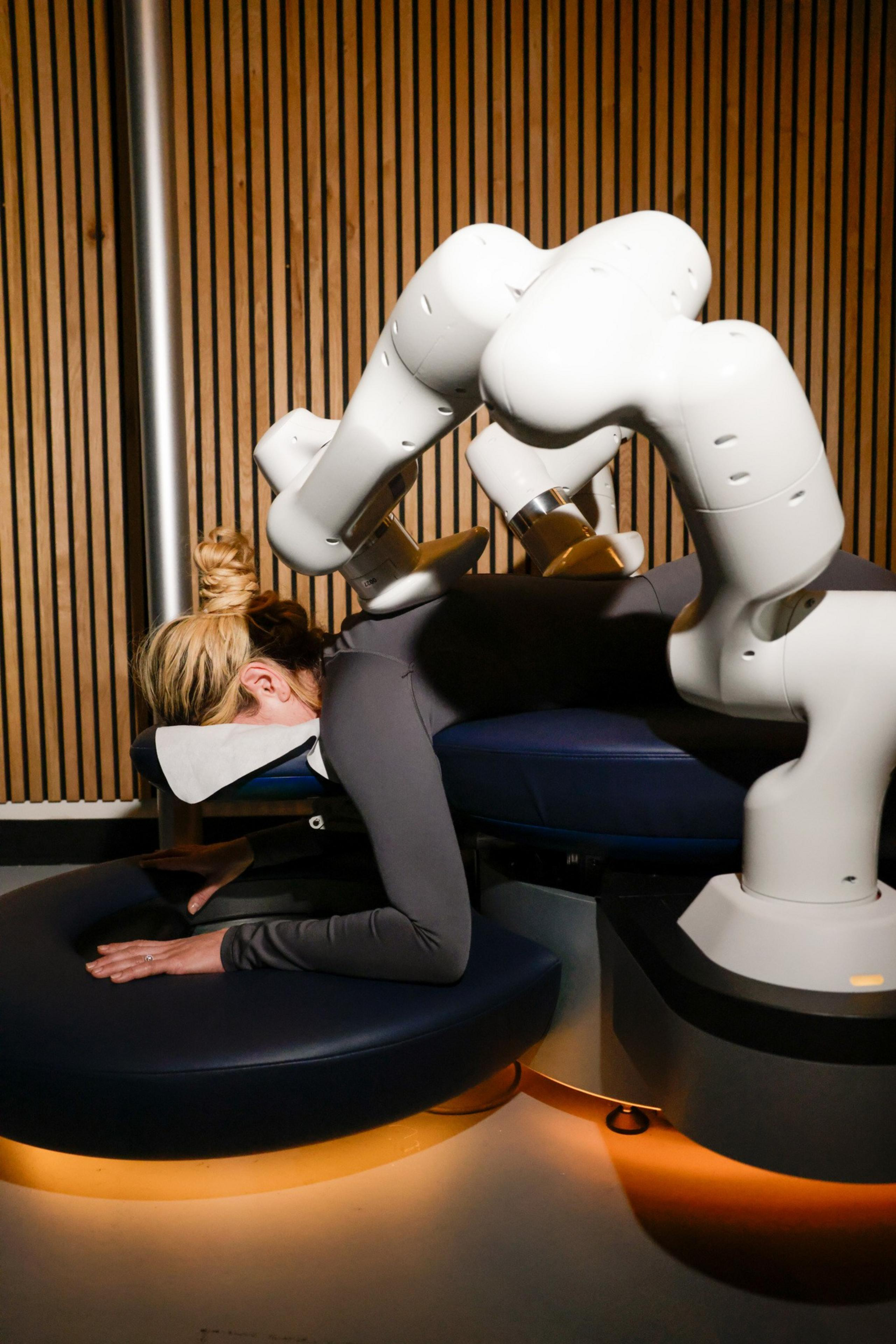 A person is lying face down on a massage table, with a robot arm hovering over their back, in a room with wooden paneling.