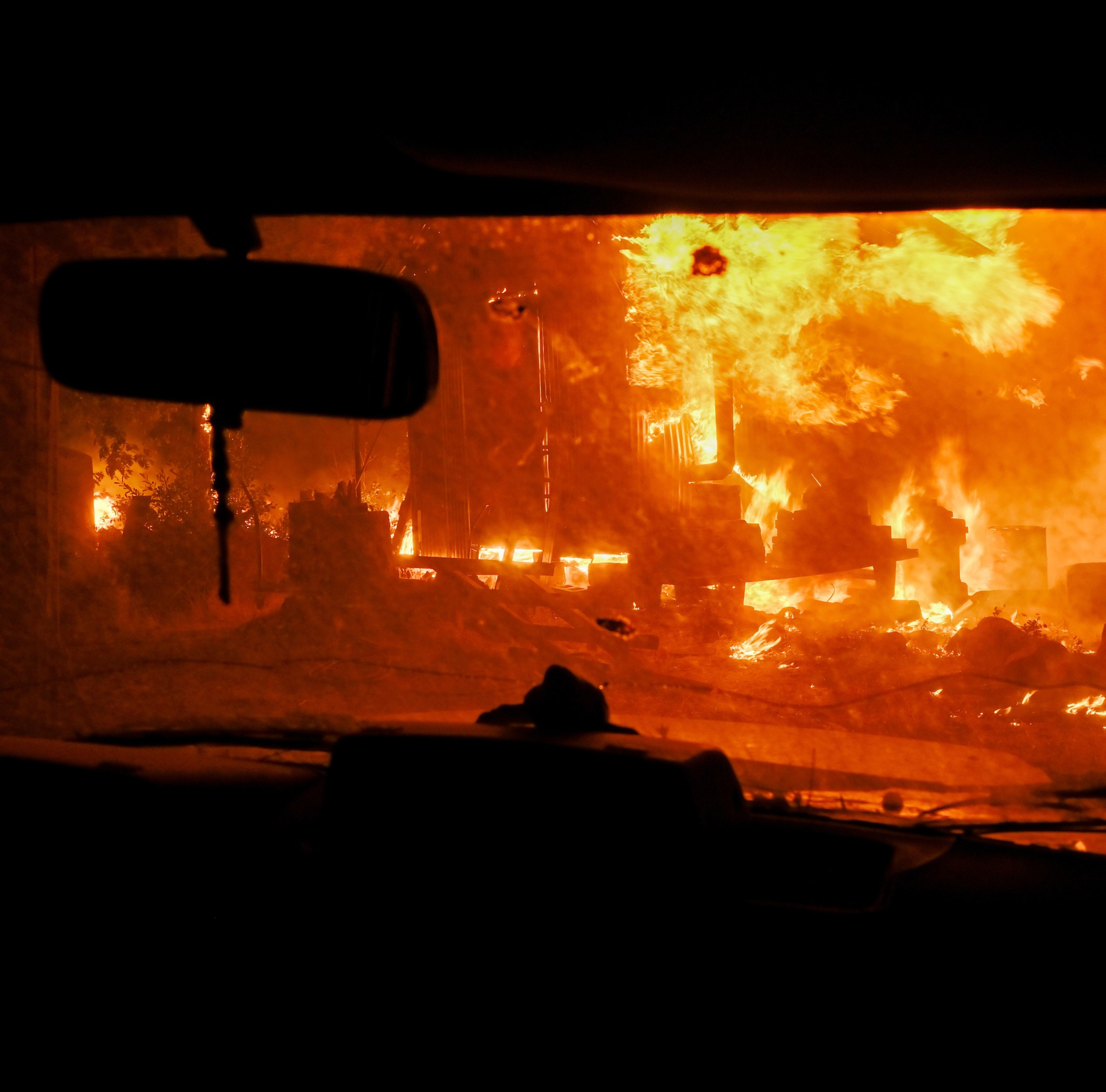 The image shows a massive fire viewed from inside a vehicle. Flames engulf structures and debris outside the windshield, casting an intense orange glow.