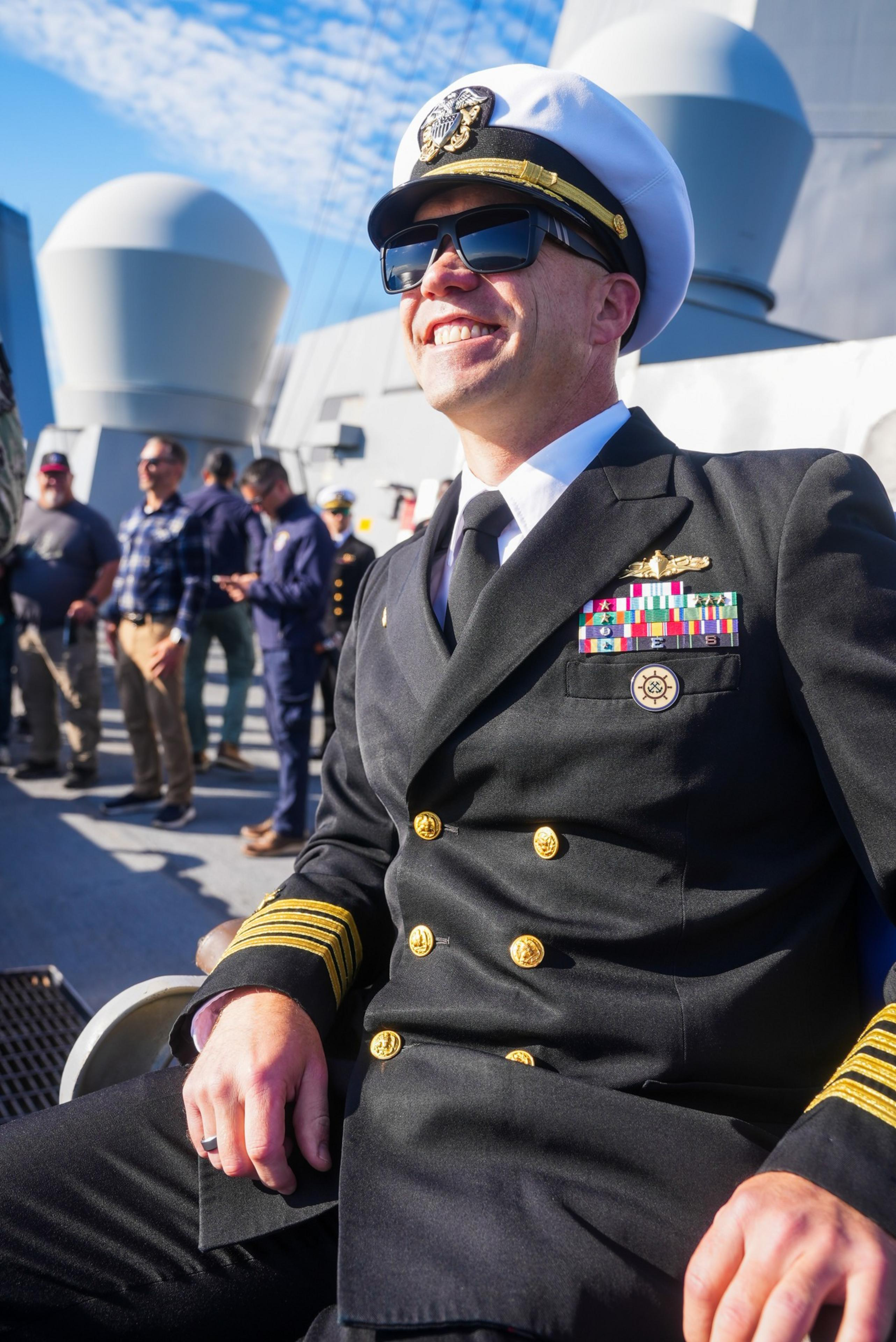 A naval officer in a double-breasted uniform, adorned with colorful ribbons, smiles broadly on a ship deck. People are gathered in the background.