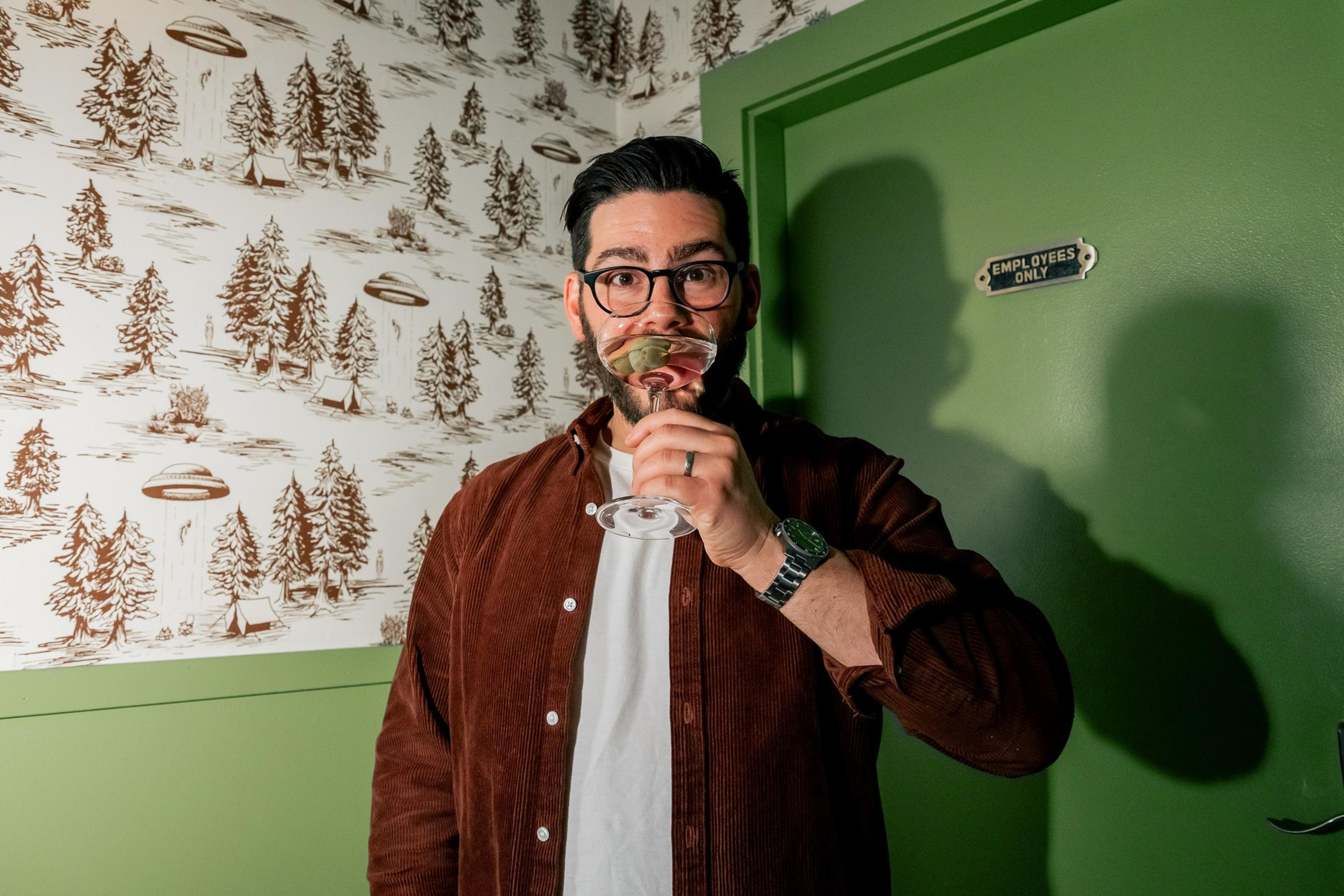 A person stands in front of a green door labeled "Employees Only," sipping from a glass. The background wallpaper shows trees and UFOs.
