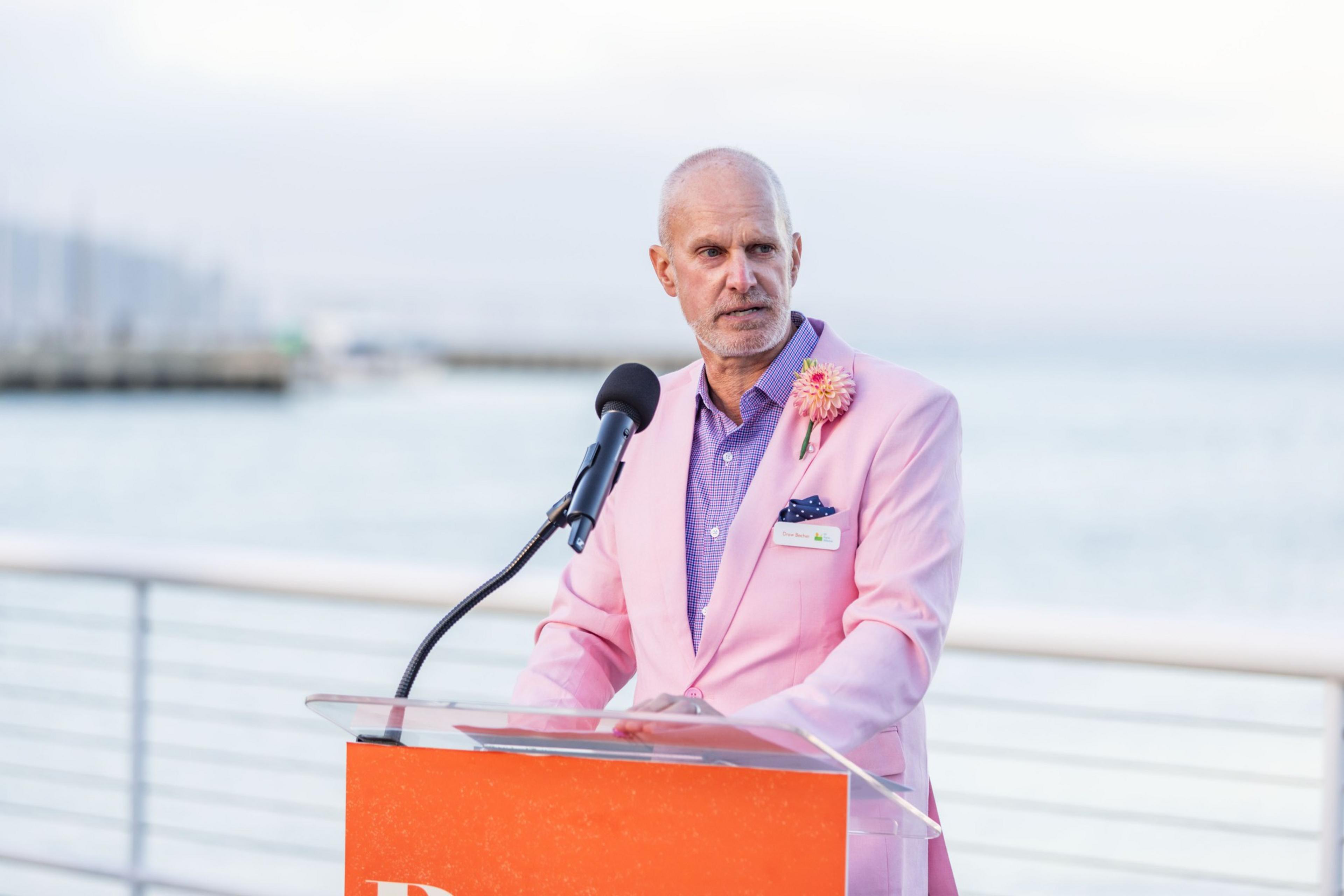 A man in a pink blazer with a flower on the lapel stands at a podium with a microphone. The background shows a blurred waterfront scene.