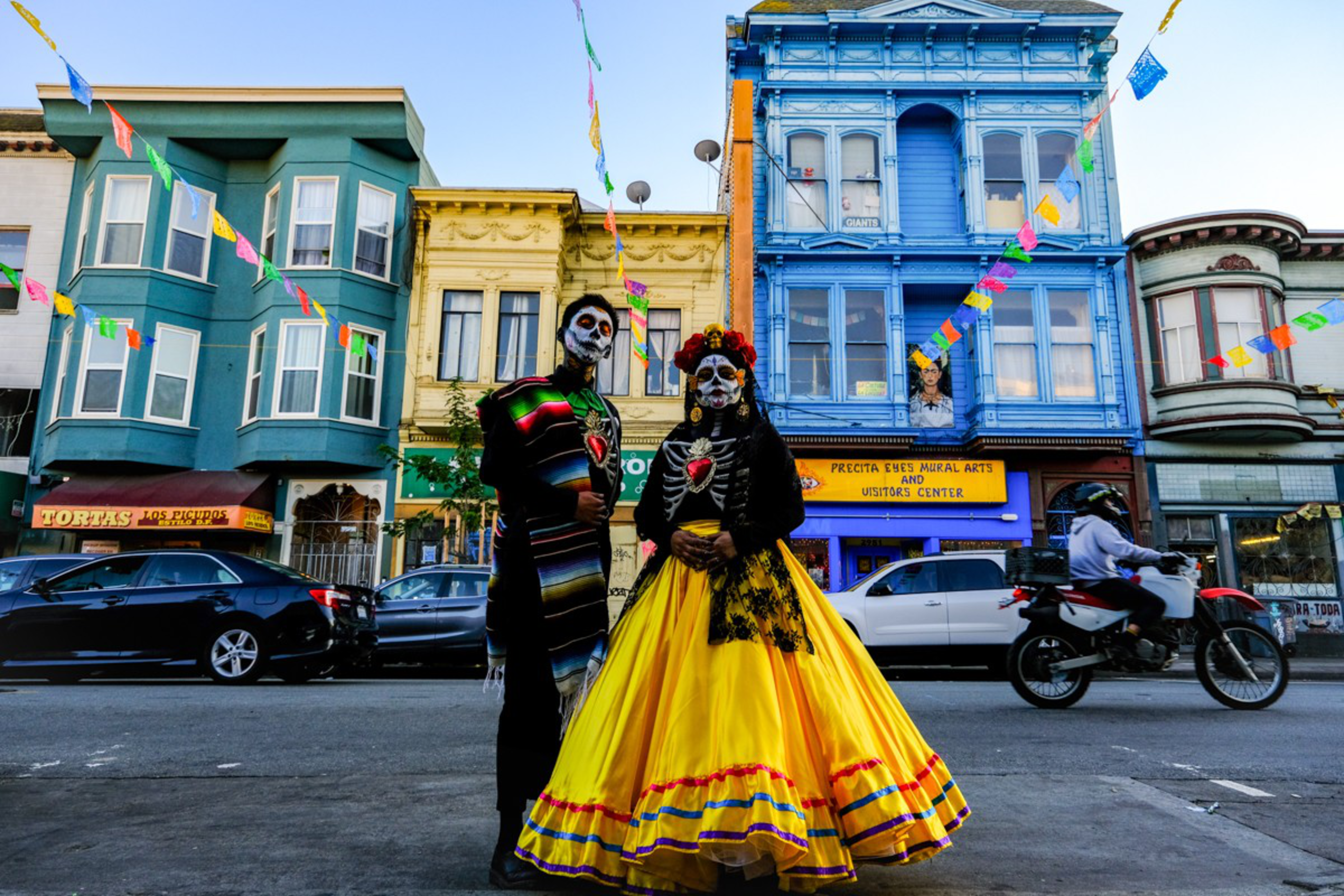 Two people in colorful Day of the Dead costumes and skull face paint stand on a street with vibrant buildings and festive flags overhead.