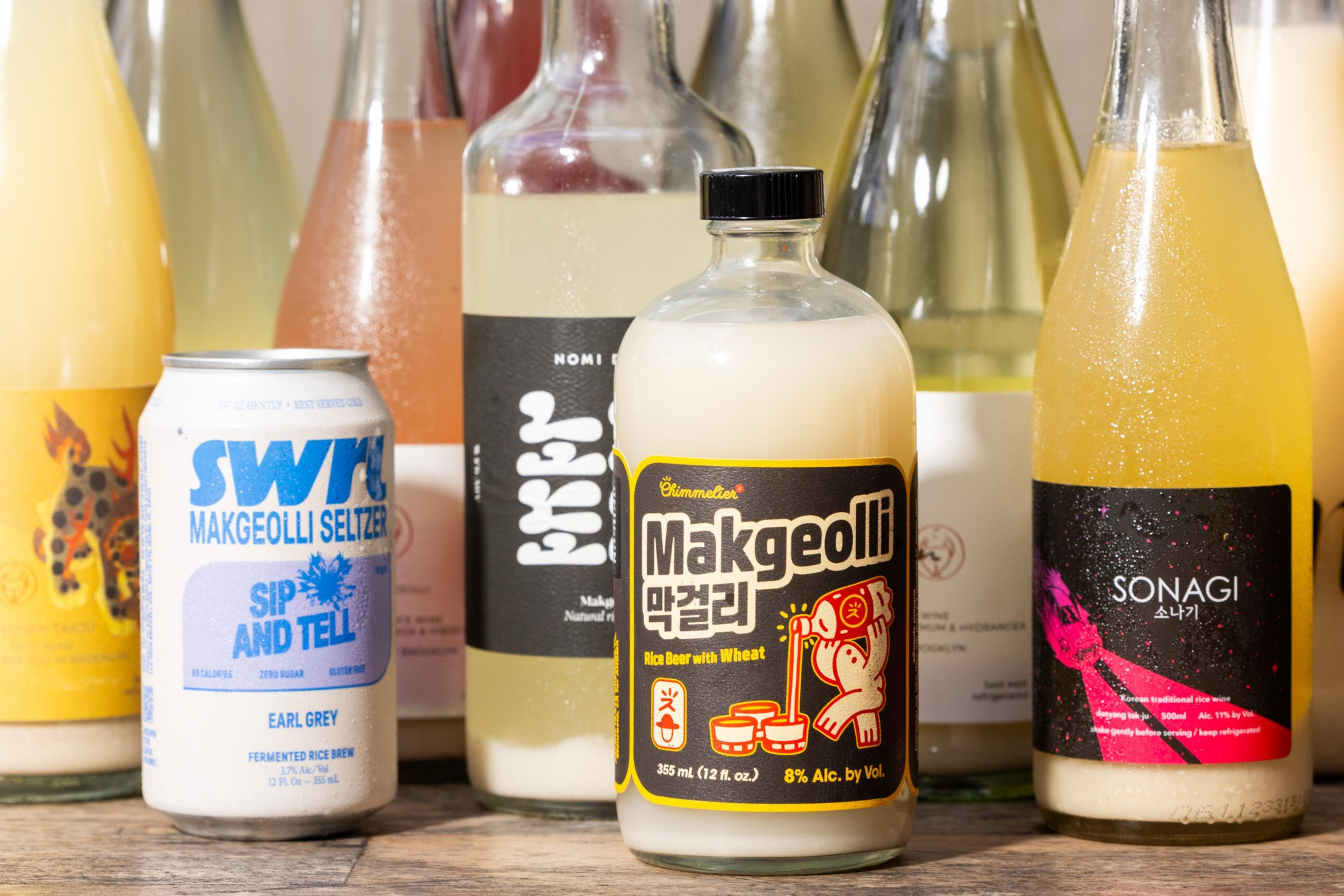 The image features a variety of makgeolli bottles and a can, with colorful labels. The central bottle is labeled &quot;Makgeolli&quot; and sits among other similarly styled drinks.