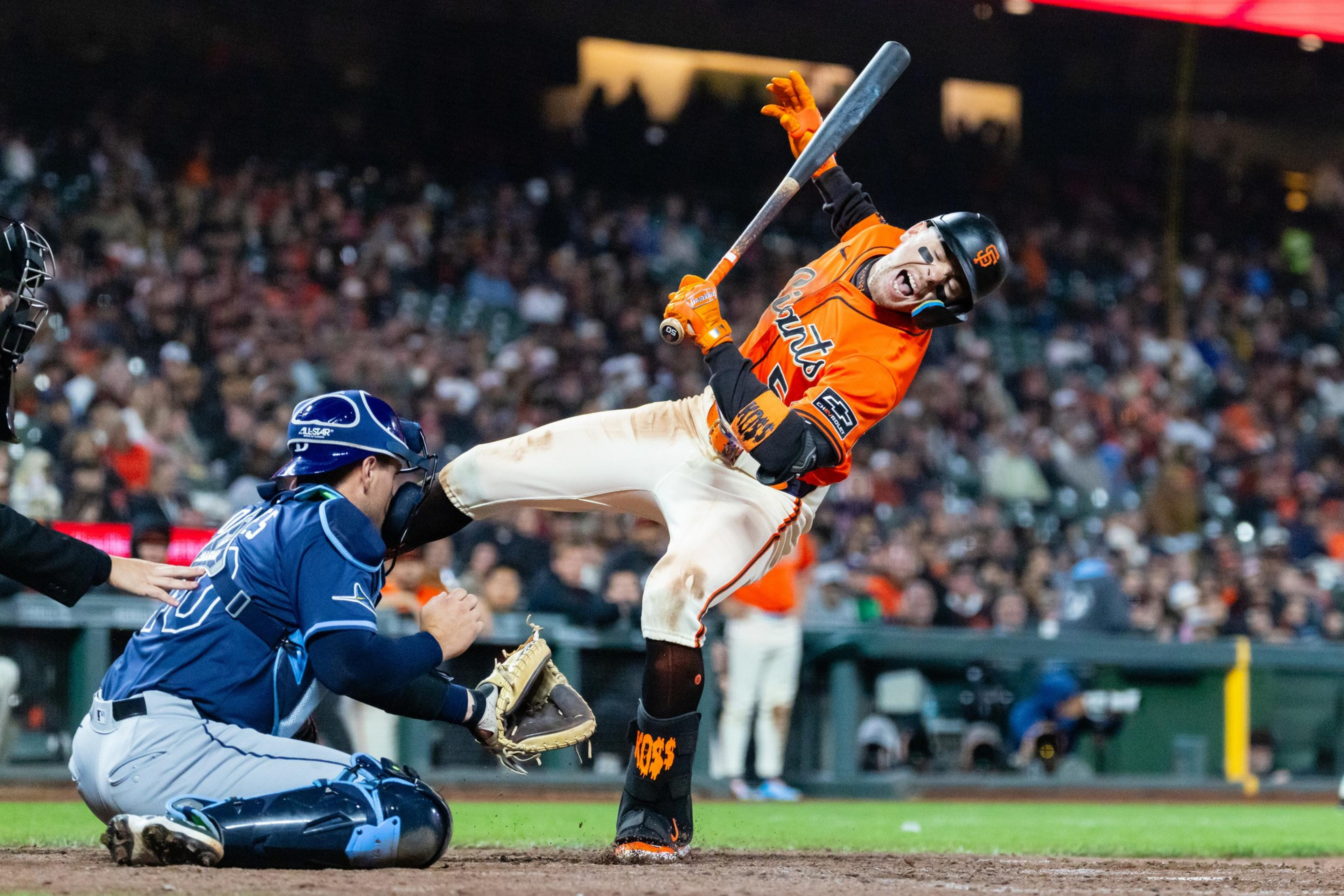 A baseball player in an orange Giants uniform is hit on the leg by a pitch as the catcher in blue gear attempts to catch the ball.
