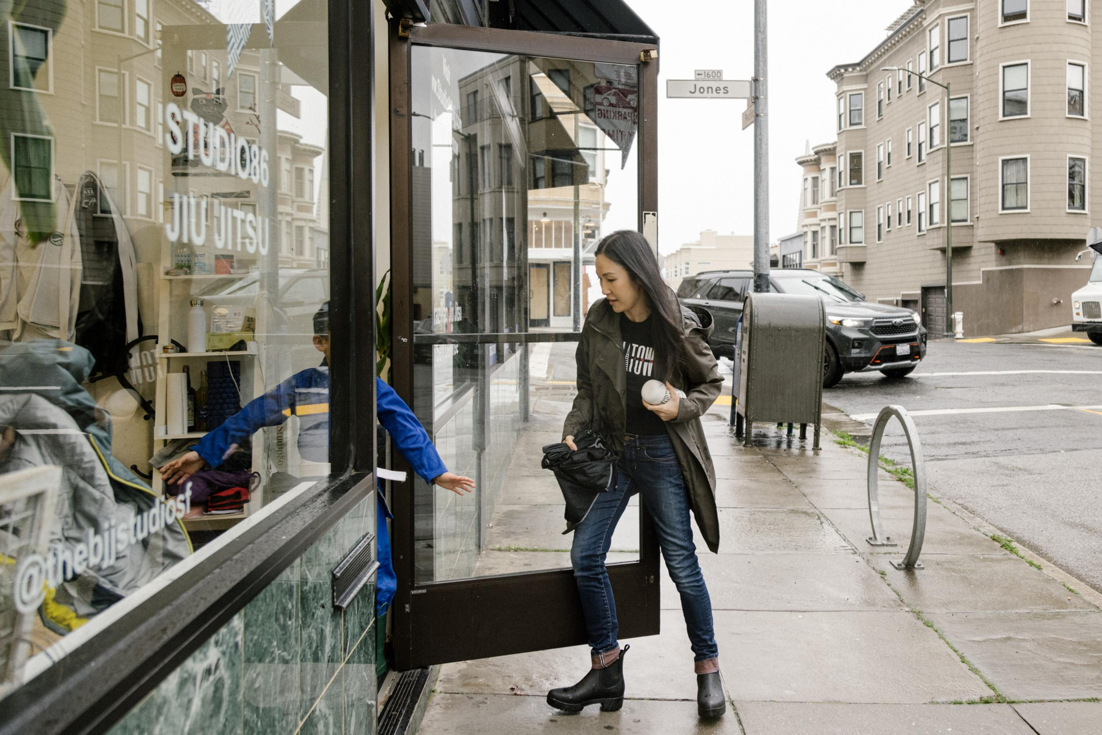 A woman exits a Jiu Jitsu studio onto a rainy street, with a child peeking from inside.