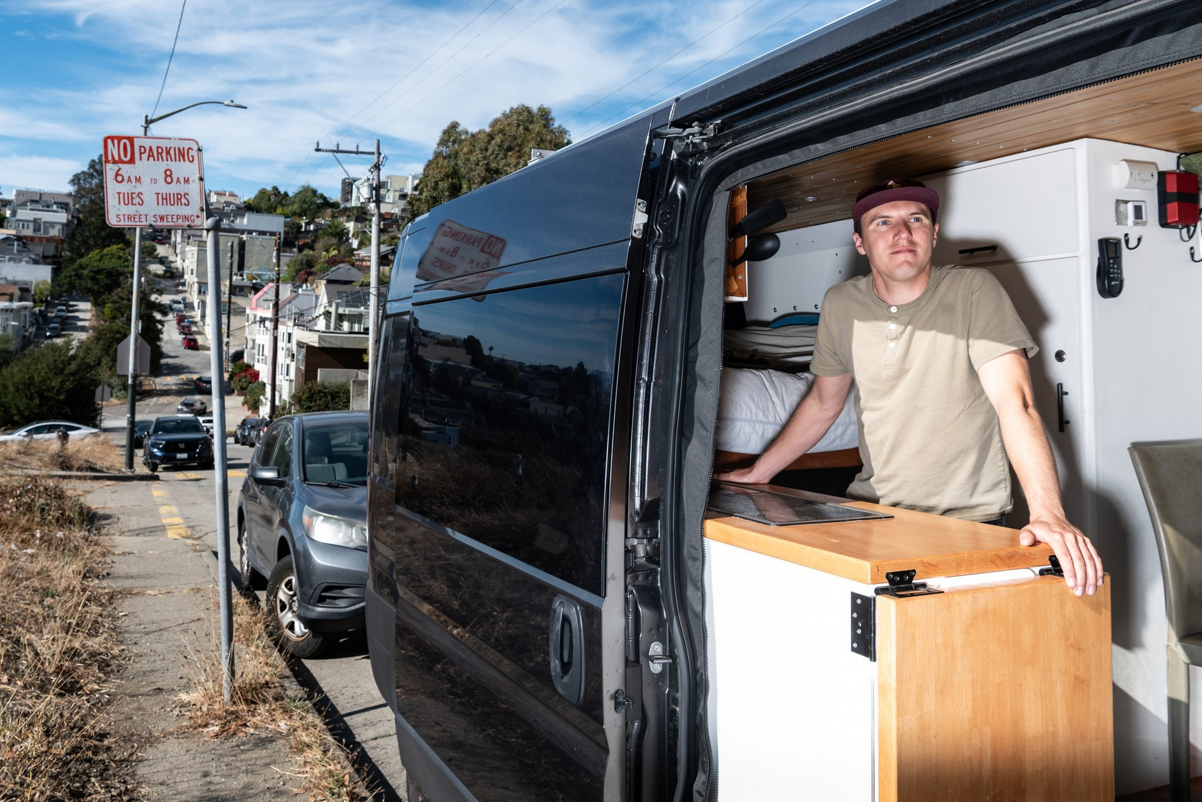 A man wearing a cap stands inside a black van with the side door open, leaning on a wooden counter in a residential neighborhood on a sunny day.