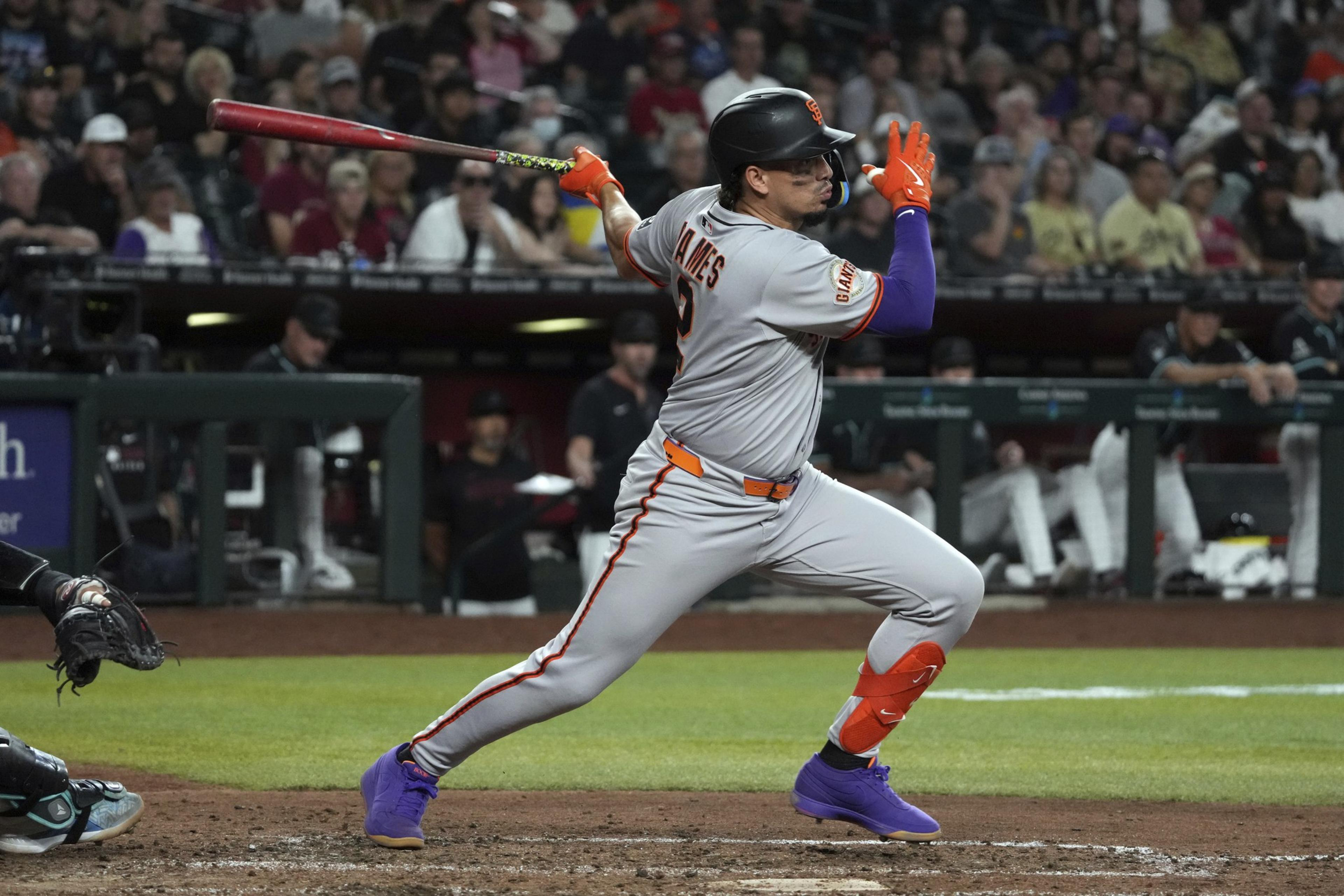 A baseball player in a gray uniform swings a bat during a game. He wears bright gloves and shoes, with an audience in the blurred background.