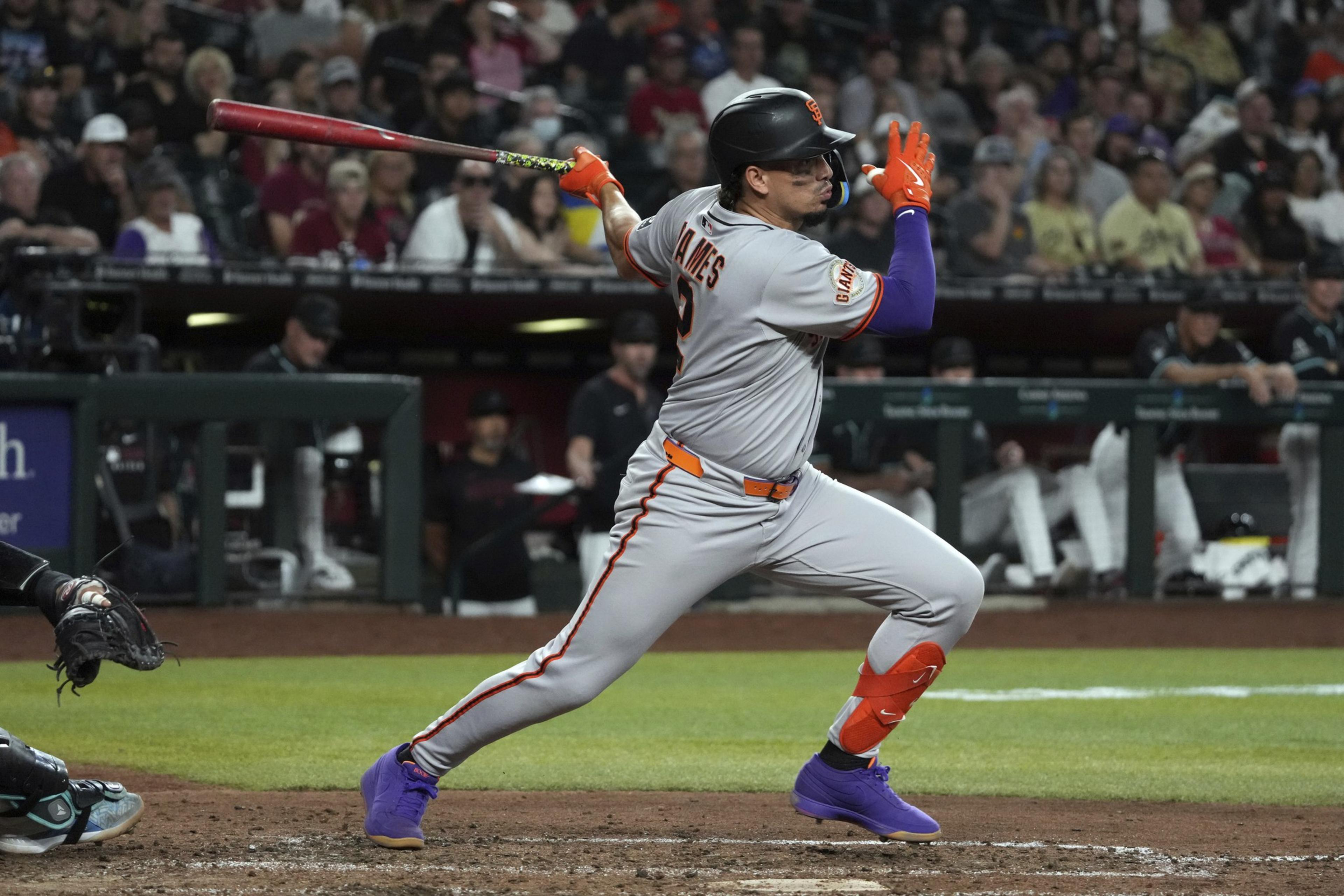 A baseball player in a gray uniform swings a bat during a game. He wears bright gloves and shoes, with an audience in the blurred background.