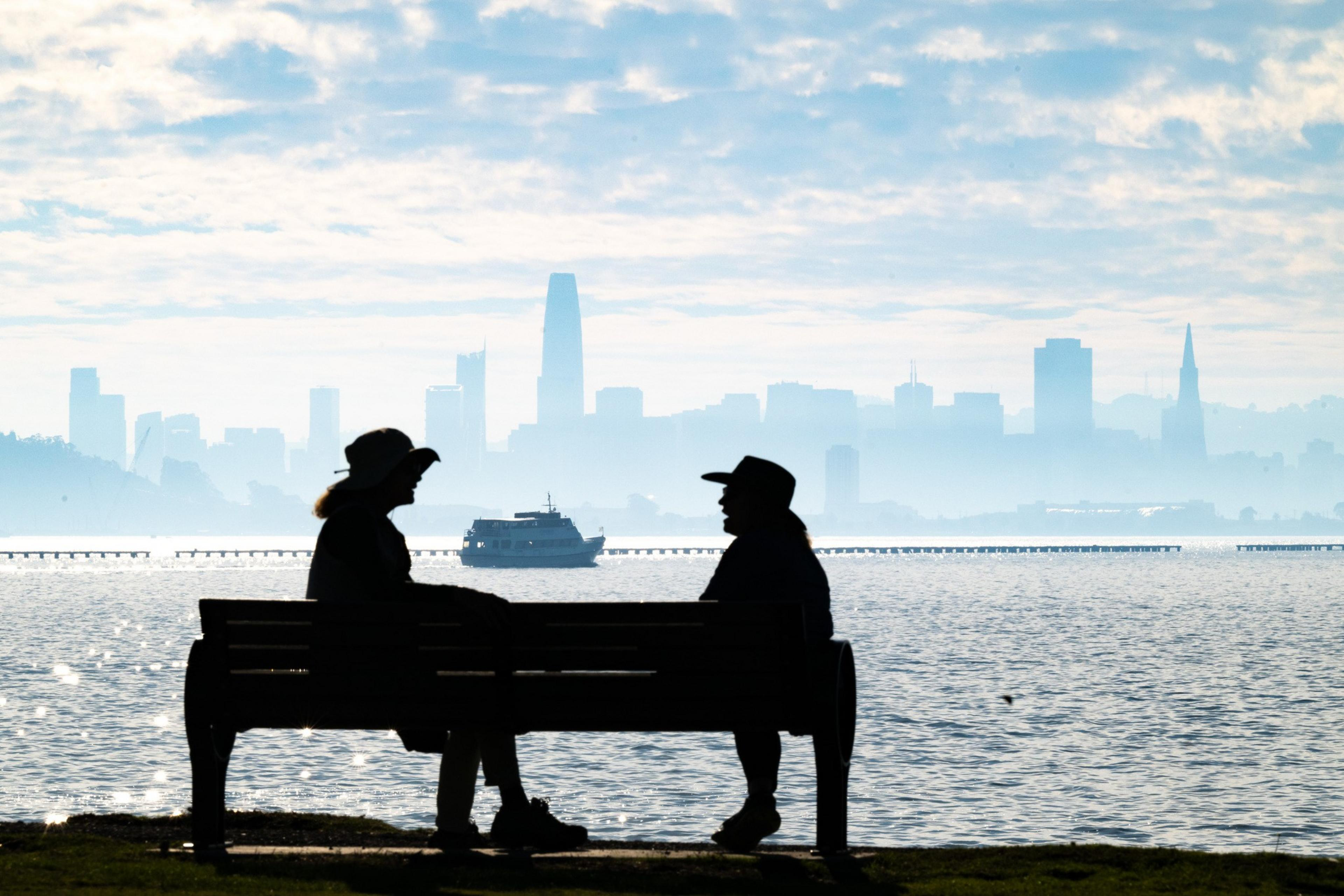 Two people in hats sit on a bench with a misty city skyline and a ferry on the water in the background.