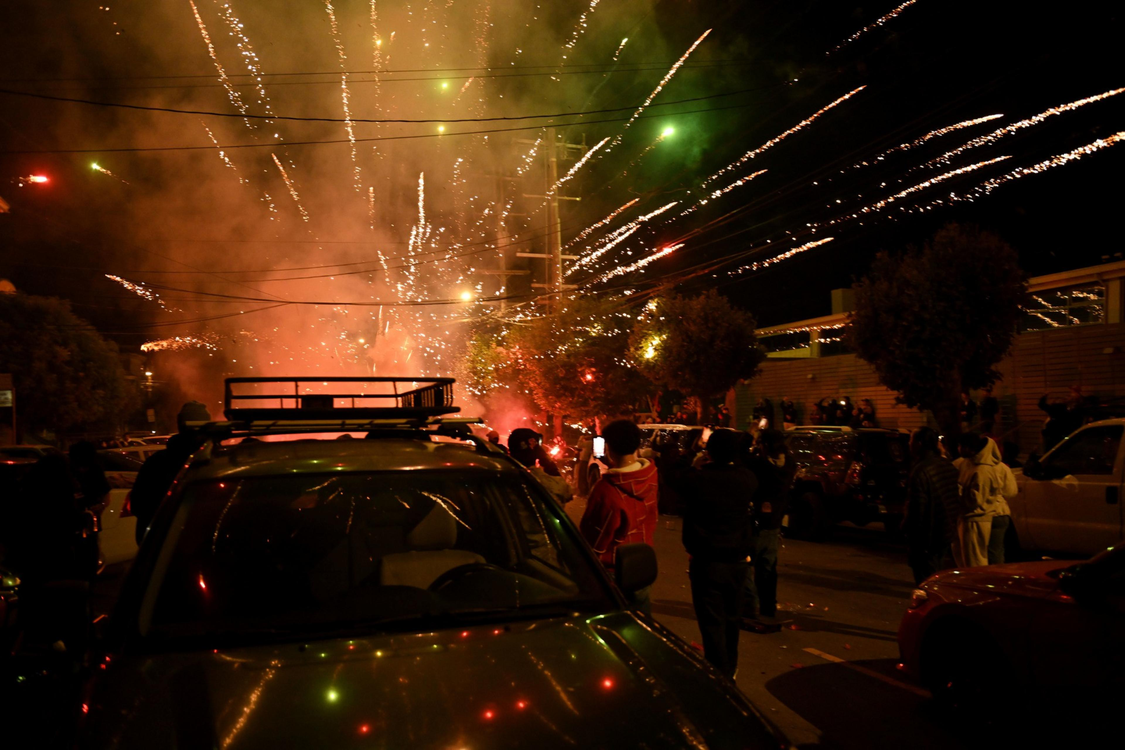 A group of people stands outside at night, looking towards fireworks in the sky. One person in a hoodie holds a phone, possibly taking a picture, while others seem engaged and curious.
