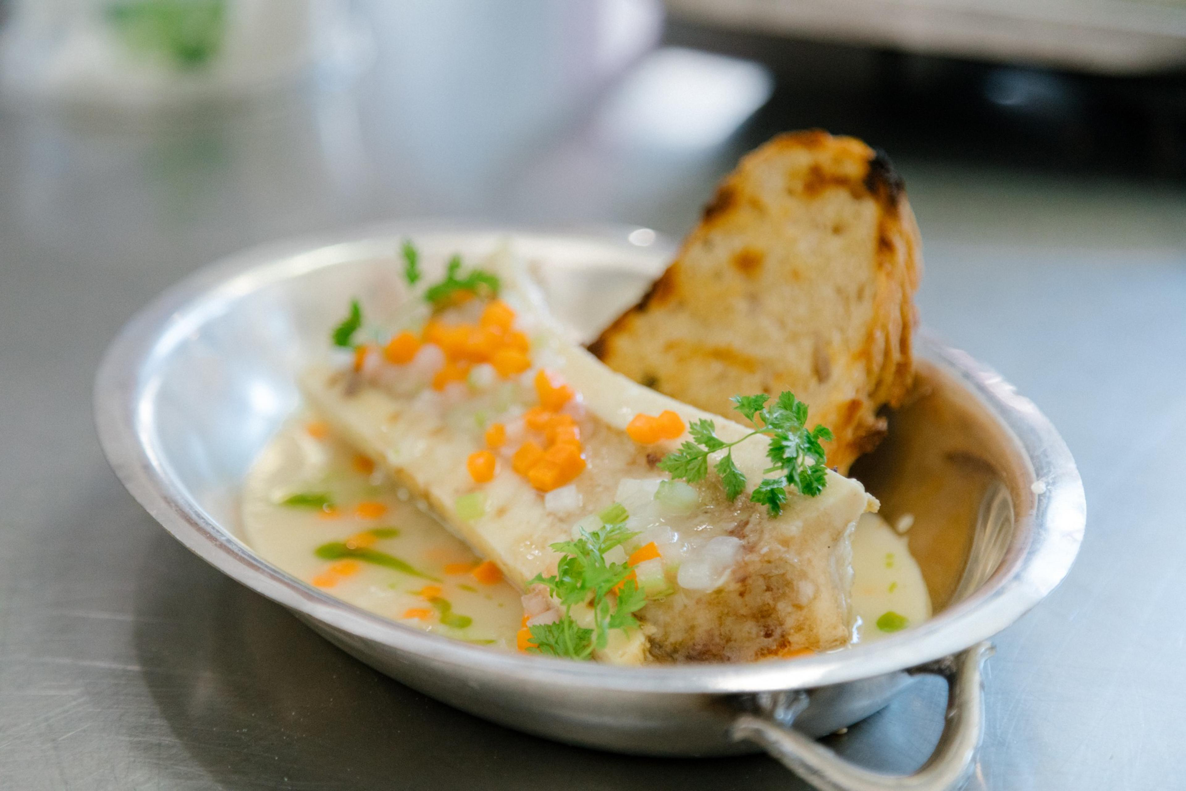 A bone marrow dish topped with herbs and diced carrots sits in a shallow metal pan, accompanied by a toasted bread slice.