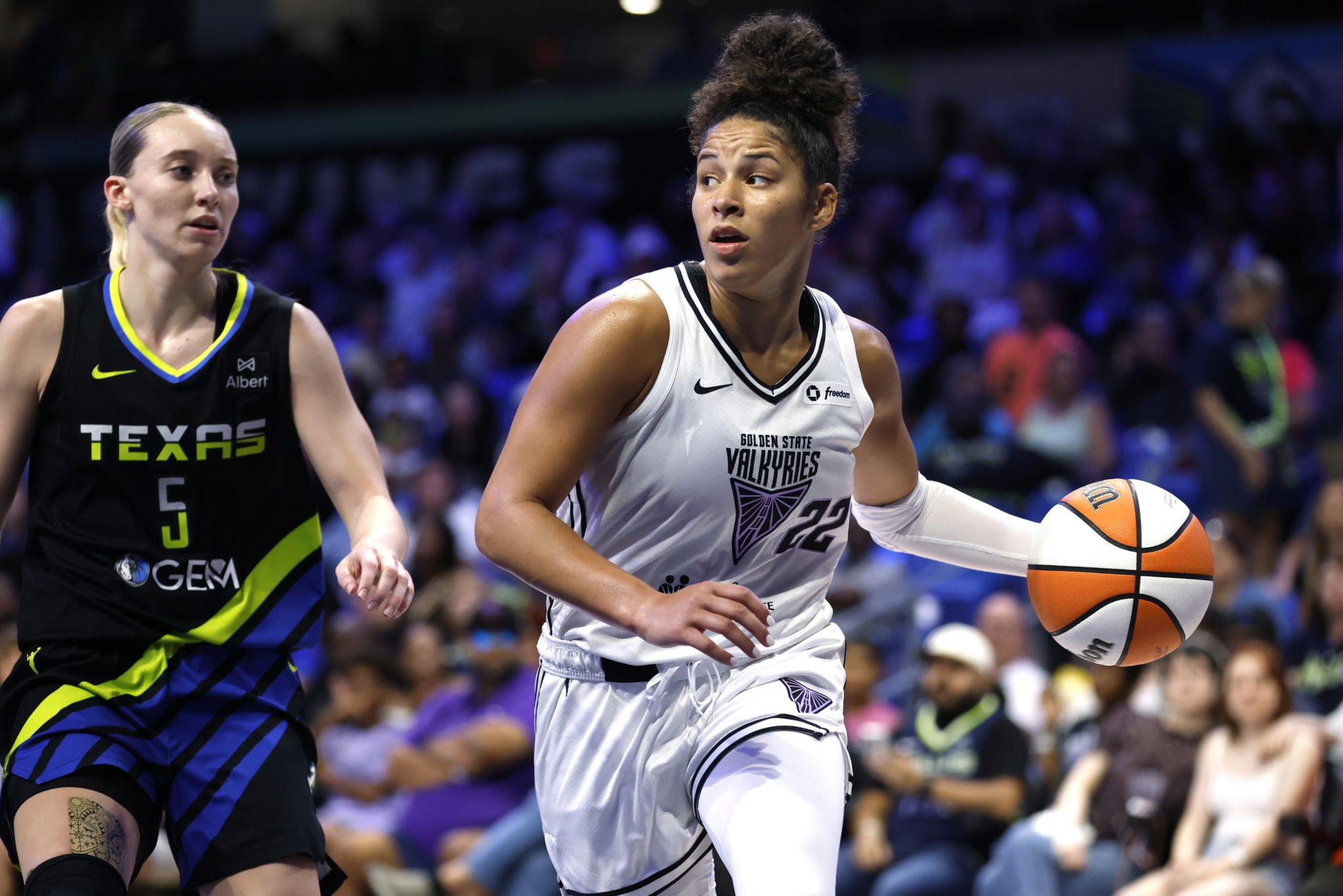 A basketball player in a white Golden State Valkyries uniform dribbles the ball, with a Texas player in a black uniform defending nearby.