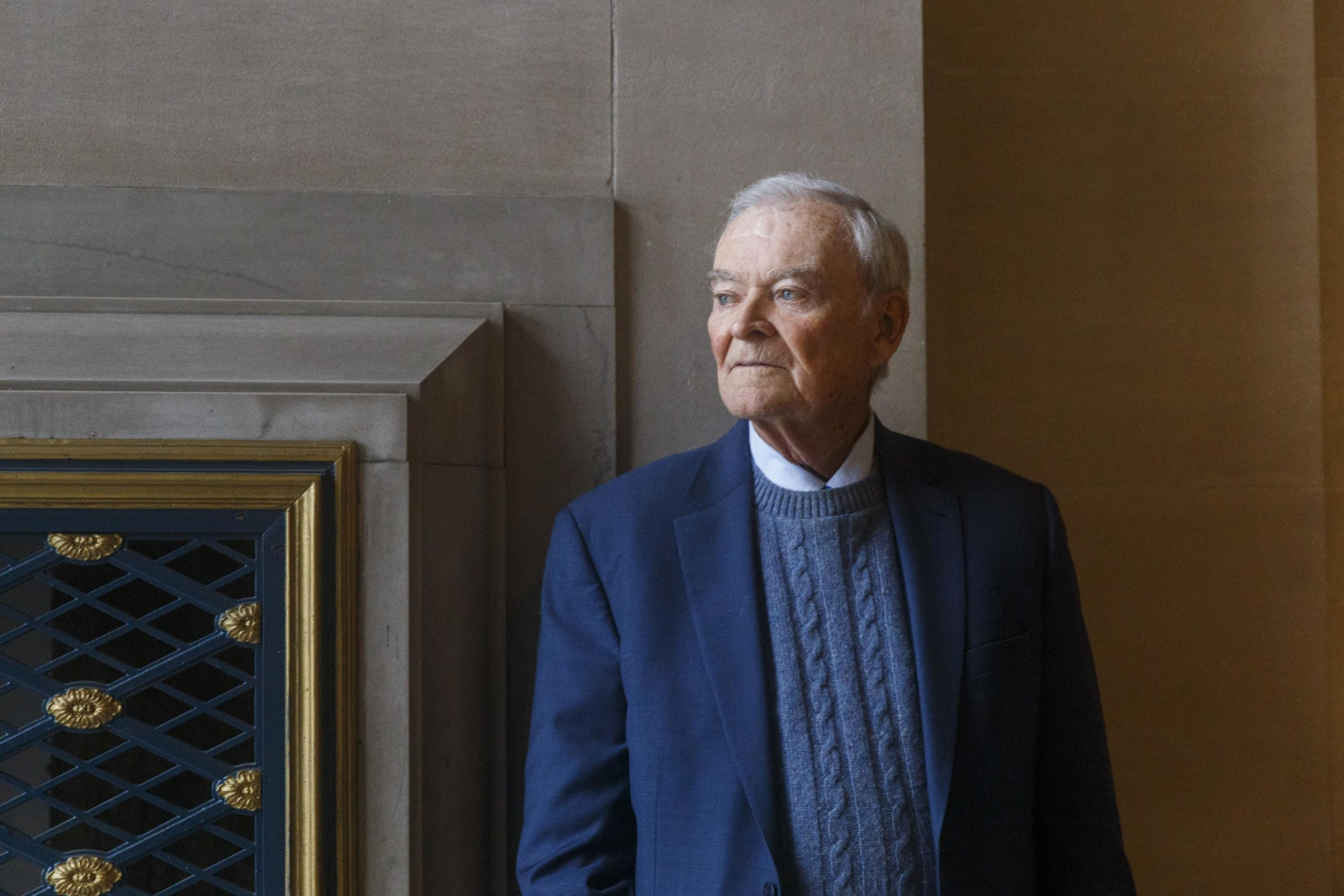 An elderly man in a blue suit and sweater stands by a wall, looking to the side pensively.