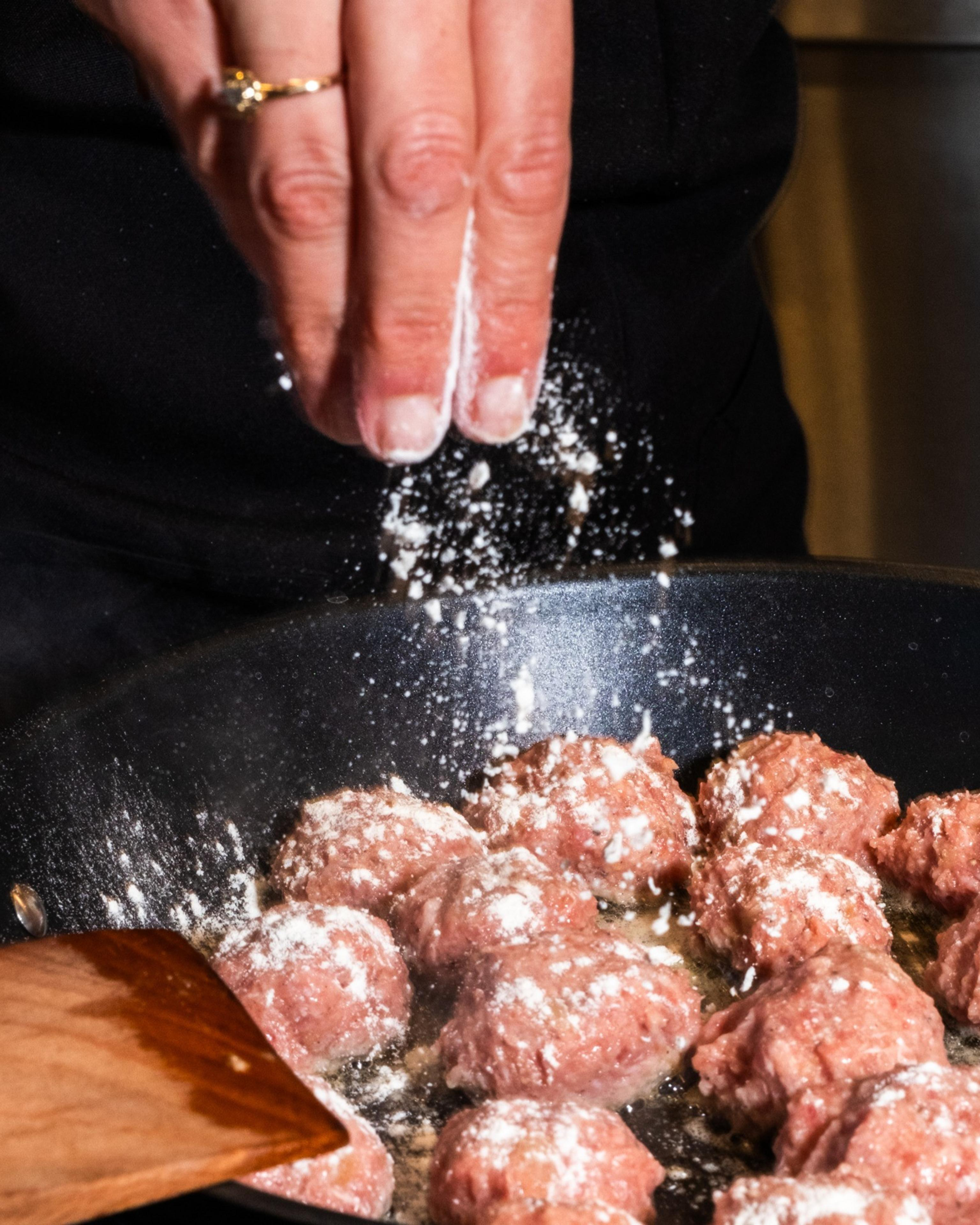 A hand sprinkles flour over meatballs sizzling in a pan, with steam rising and a wooden spatula nearby, creating a dynamic cooking scene.