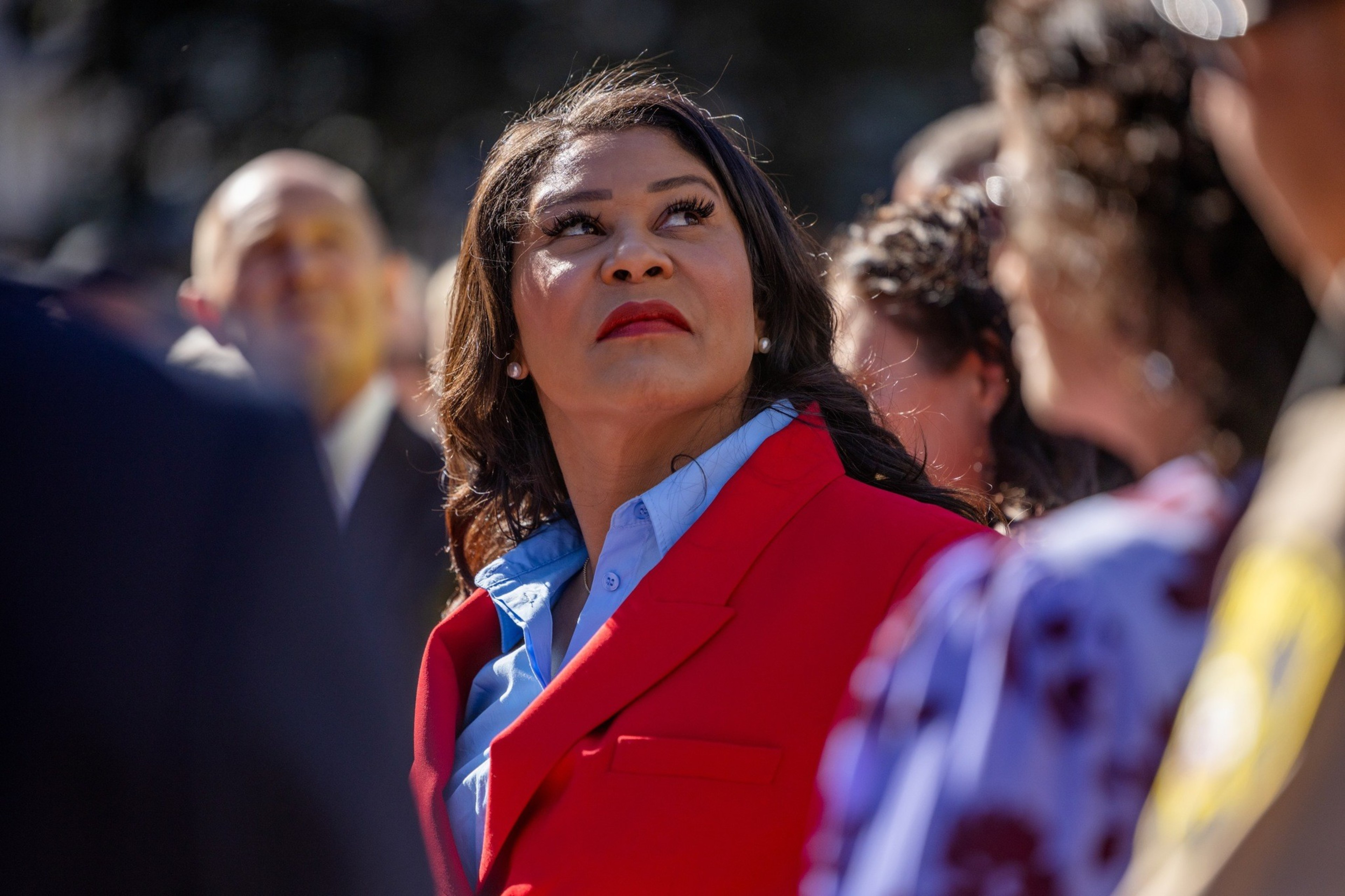 San Francisco Mayor London Breed, with a serious expression, looks towards the sky as she's surrounded by other unidentifiable people that are out of focus.