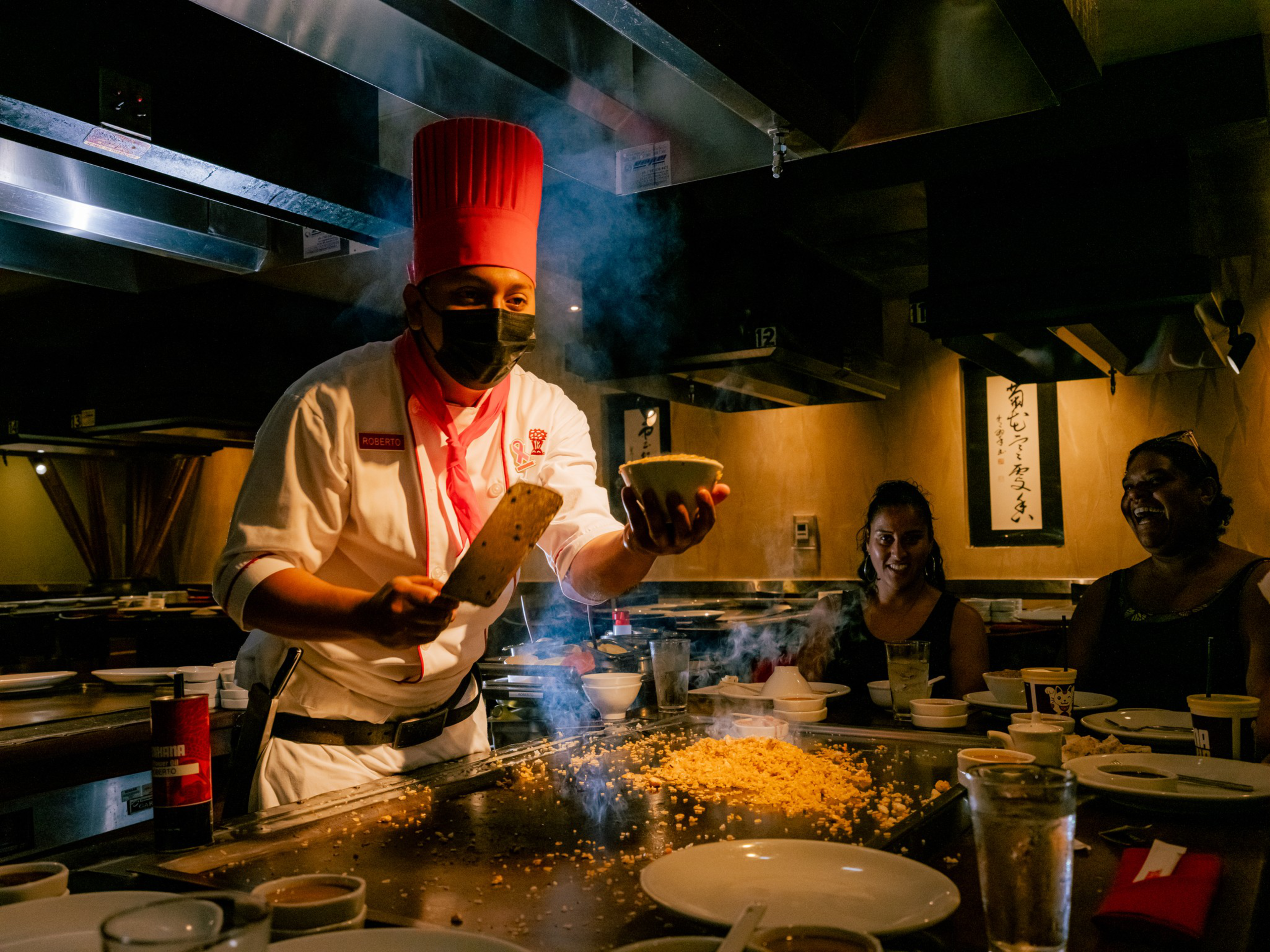 A chef wearing a red hat and mask prepares food on a hibachi grill while three diners watch and smile at a restaurant.