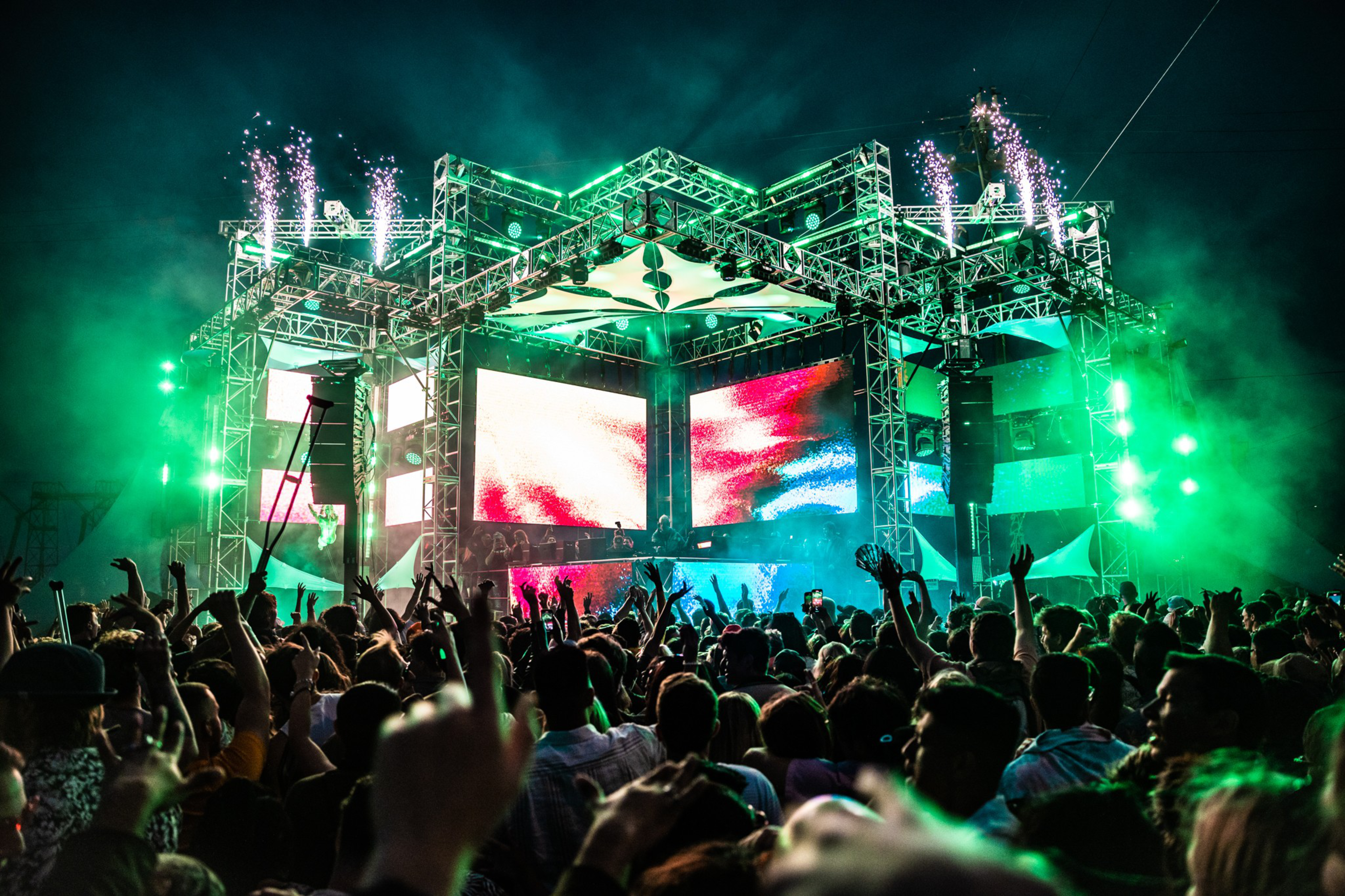 A large crowd dances and raises hands in front of a brightly lit stage with colorful screens and green lighting effects at a nighttime outdoor concert.