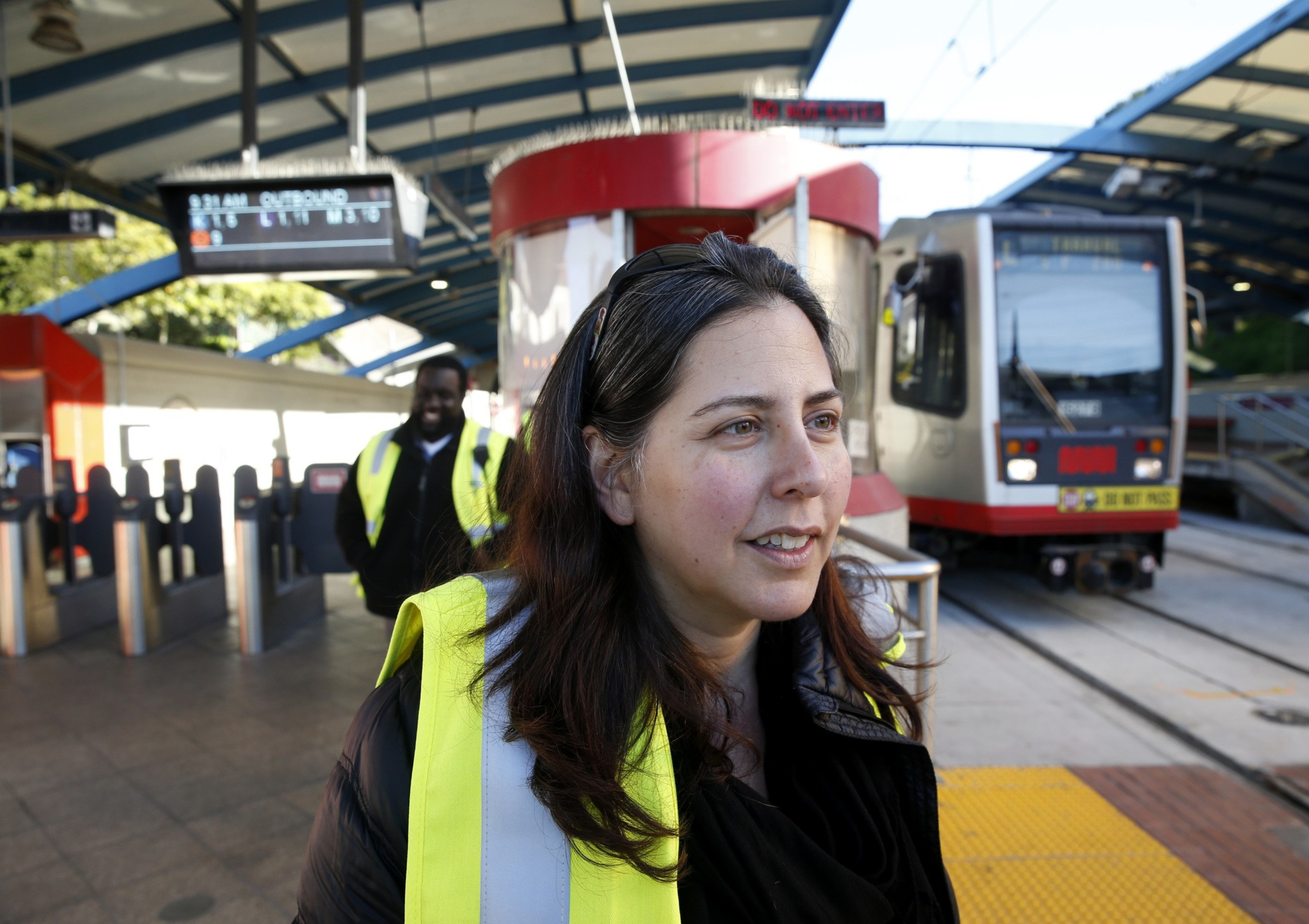 A woman in a yellow safety vest stands near a train platform, with a train visible in the background. Another person in a vest is behind her, near a turnstile area.