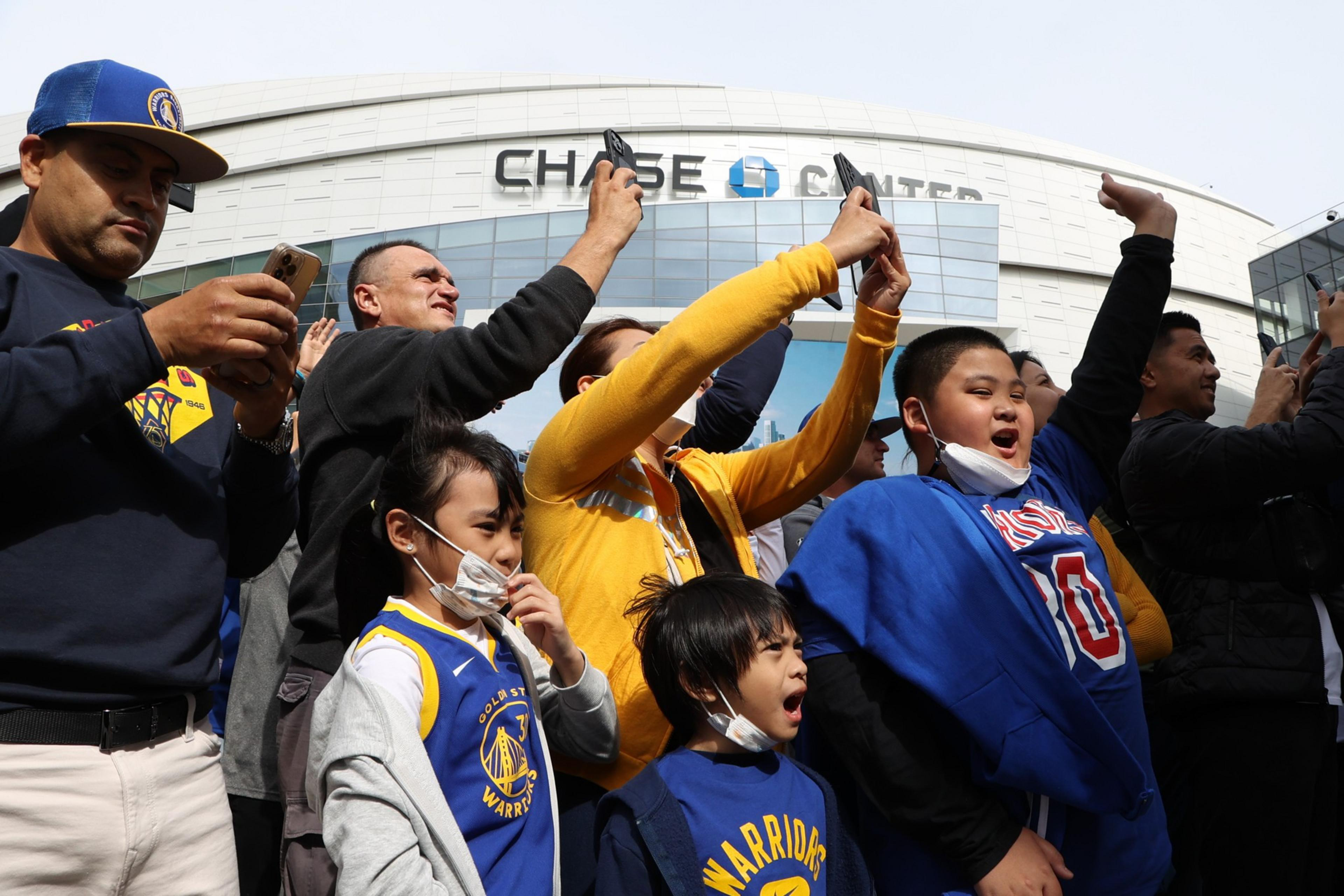 A crowd of enthusiastic people, many wearing Warriors gear, stand outside Chase Center, some holding phones, with a sense of excitement and anticipation.