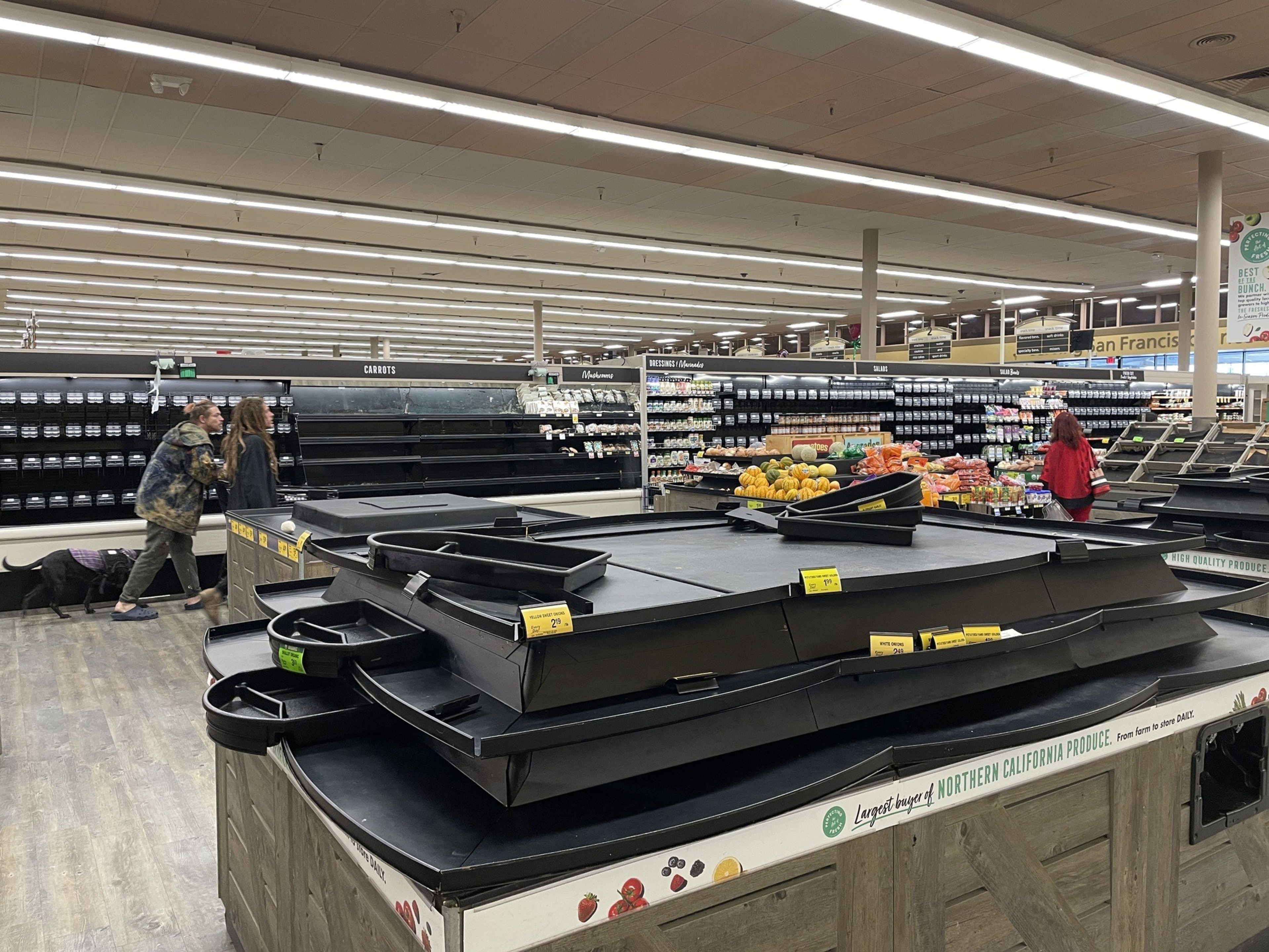 A mostly empty grocery store produce section with sparse items. Various bins are bare, and a few shoppers and scattered vegetables are seen in the background.