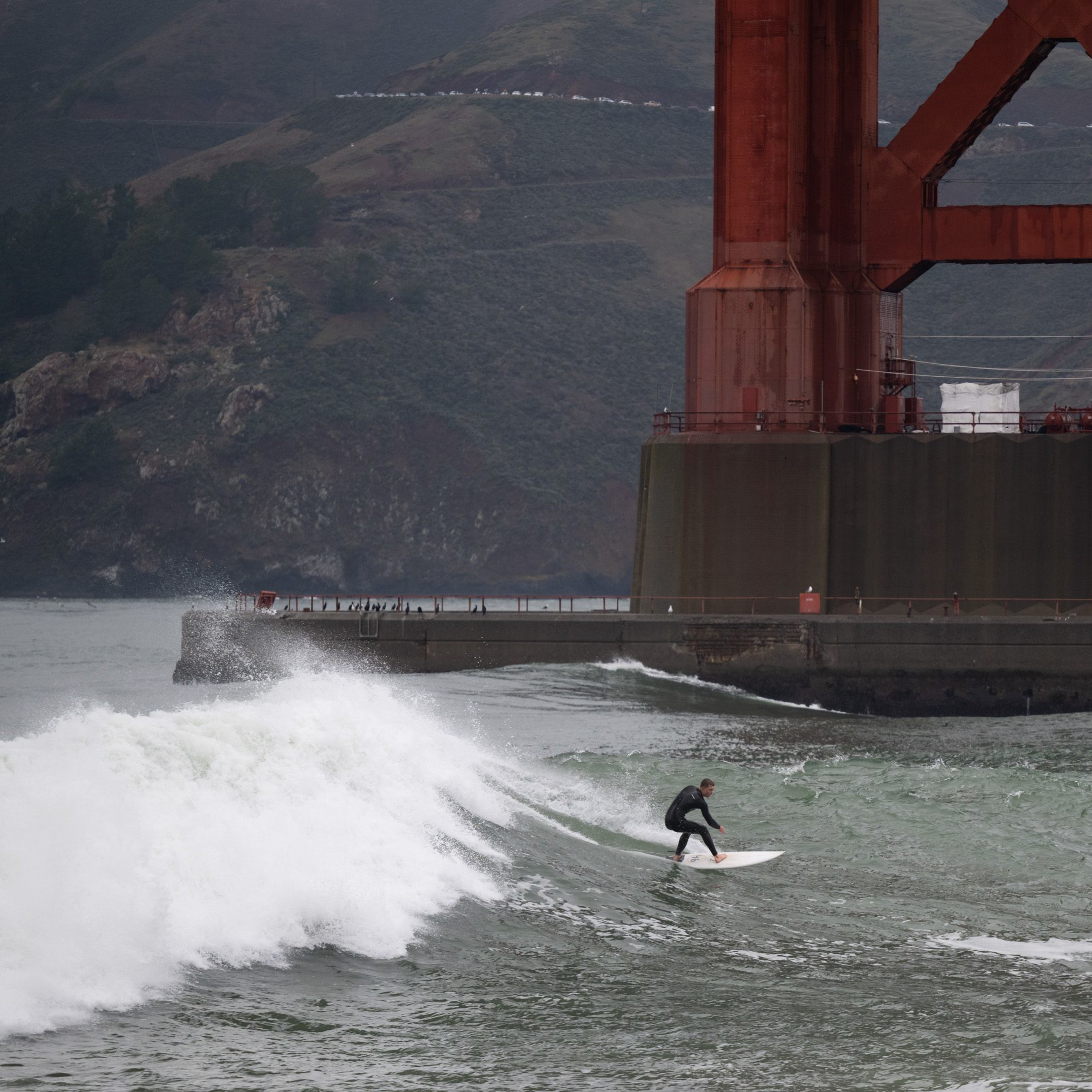 A surfer rides a wave near the base of a large, rust-colored bridge structure. In the background, there are hilly, grassy landscapes under an overcast sky.