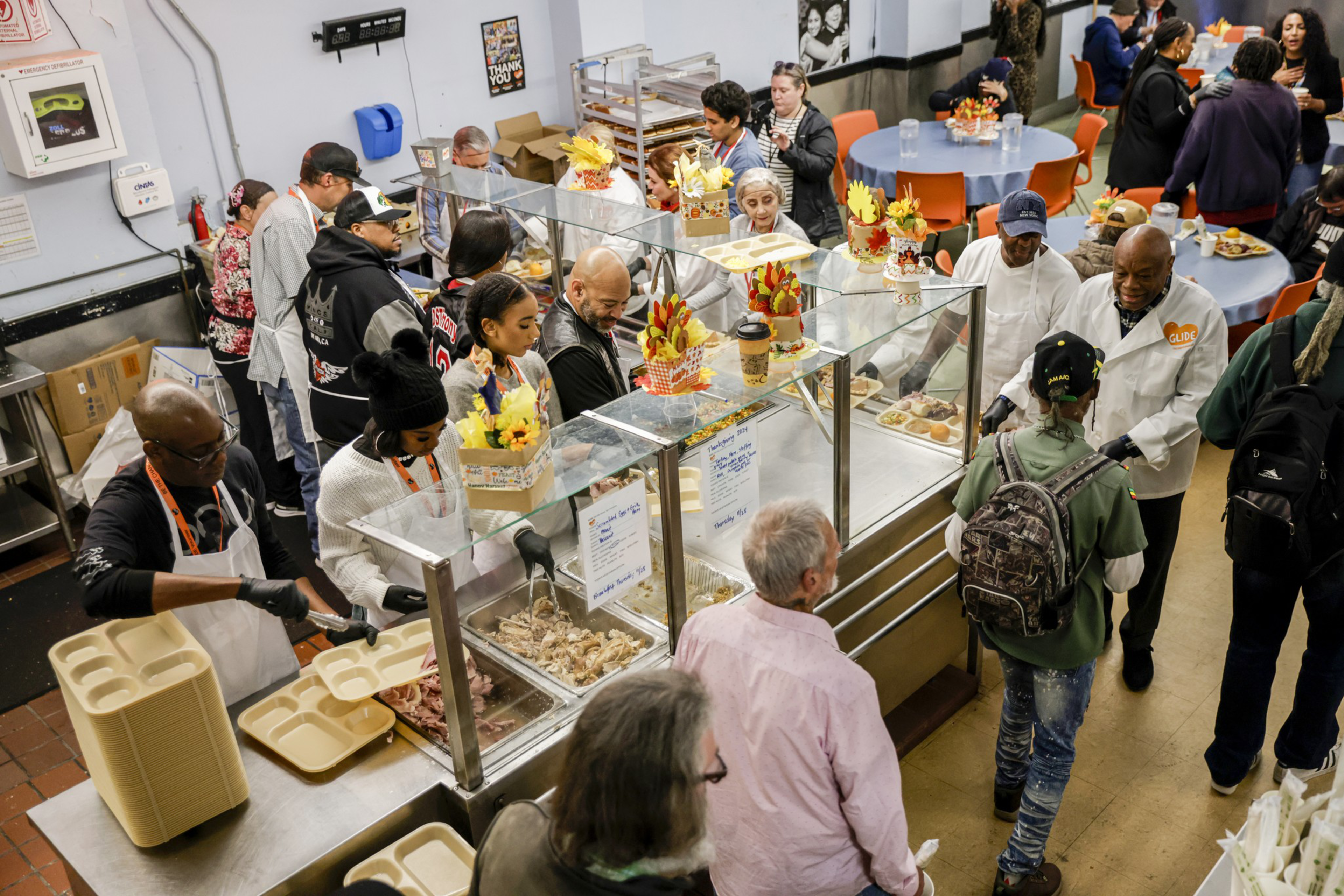 People stand in line and volunteers serve food behind a counter in a busy community kitchen or soup kitchen setting.