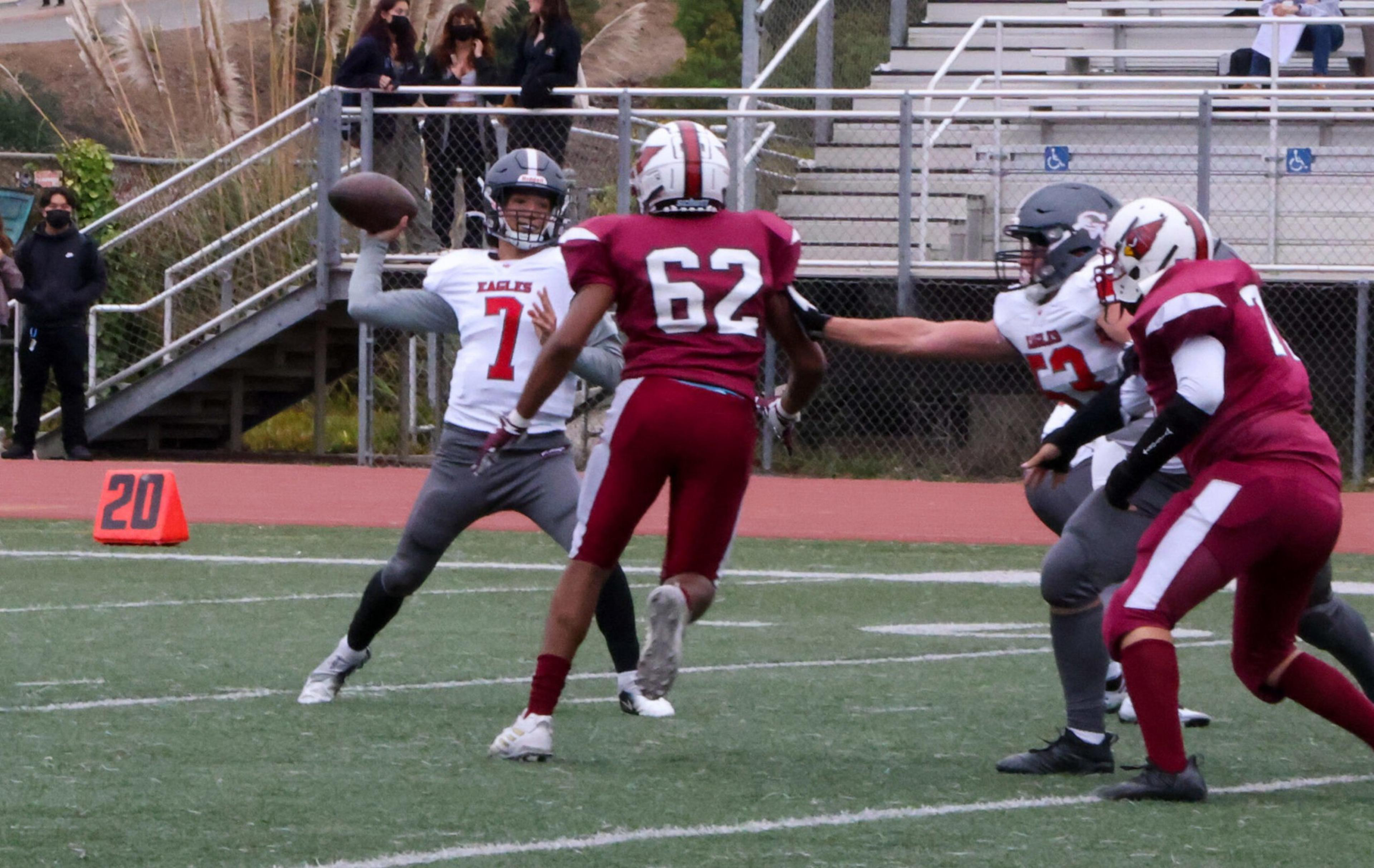 A football quarterback in a white and gray uniform prepares to throw the ball while a defender in a red uniform rushes toward him on a green field.