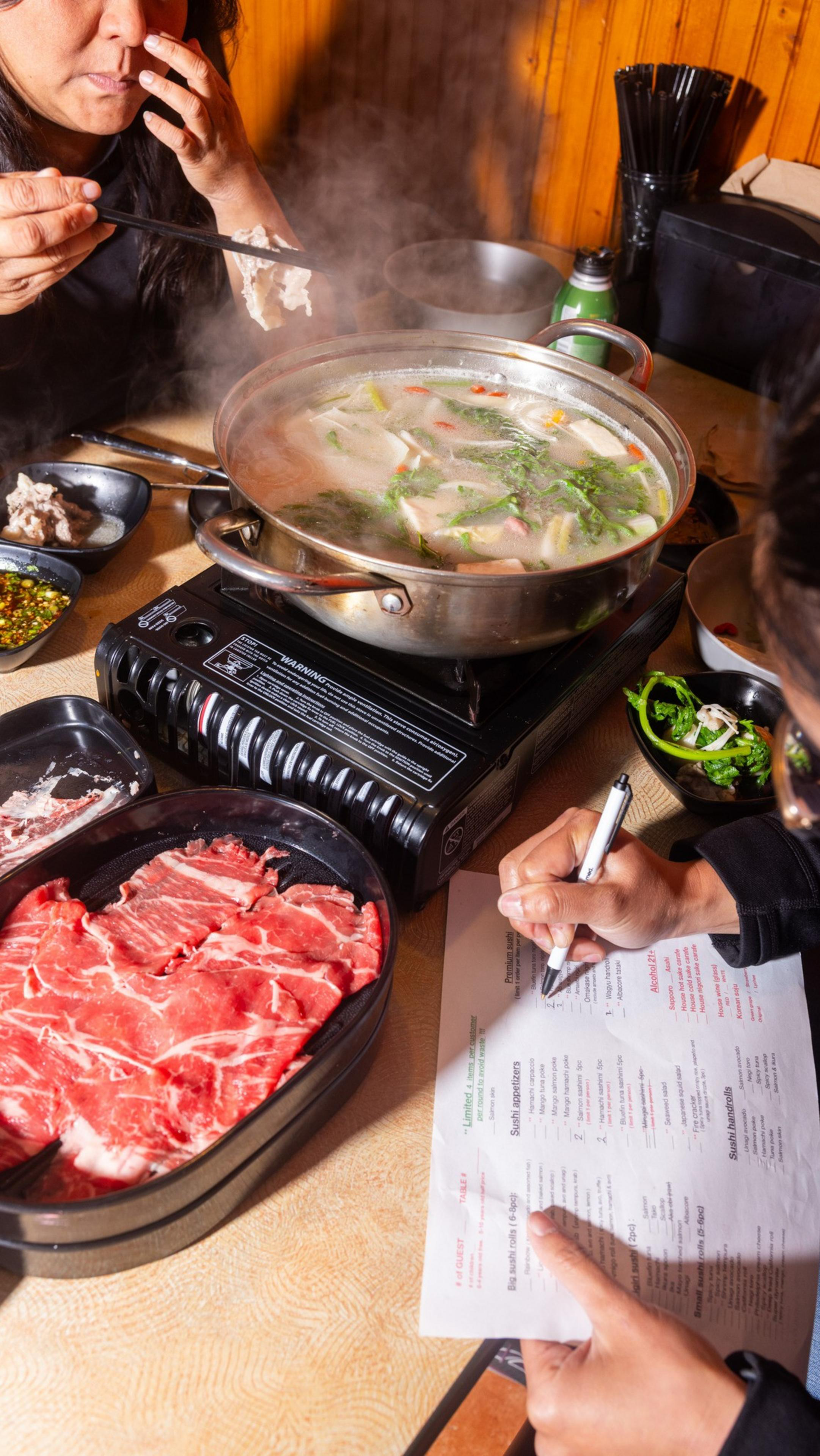 A person eats from a steaming hot pot on a table with raw meat and greens, writing on a menu. Another person holds chopsticks nearby.