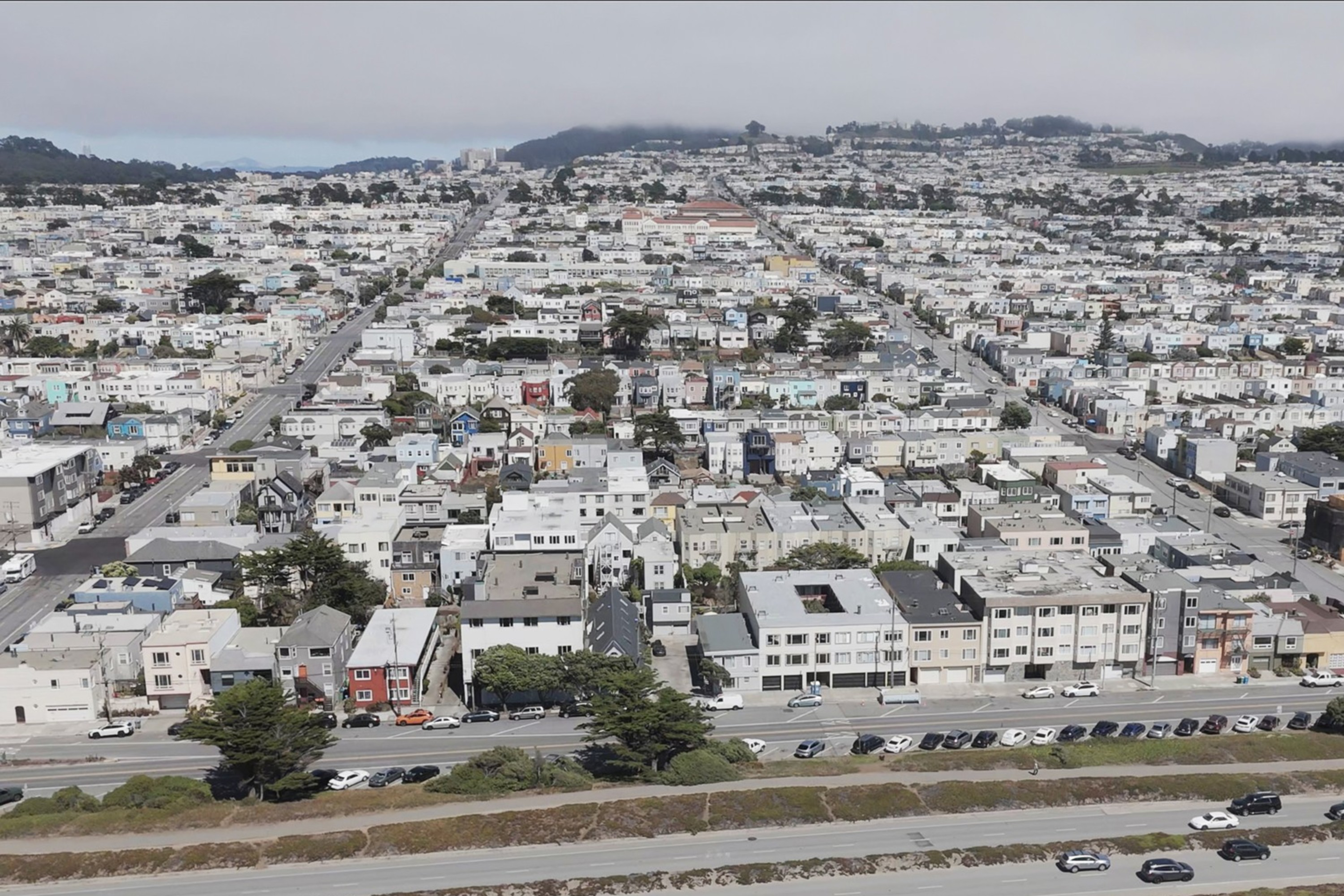 The image shows a densely packed suburban area with rows of houses, intersecting streets, and a highway with cars in the foreground. A hilly landscape is in the background.