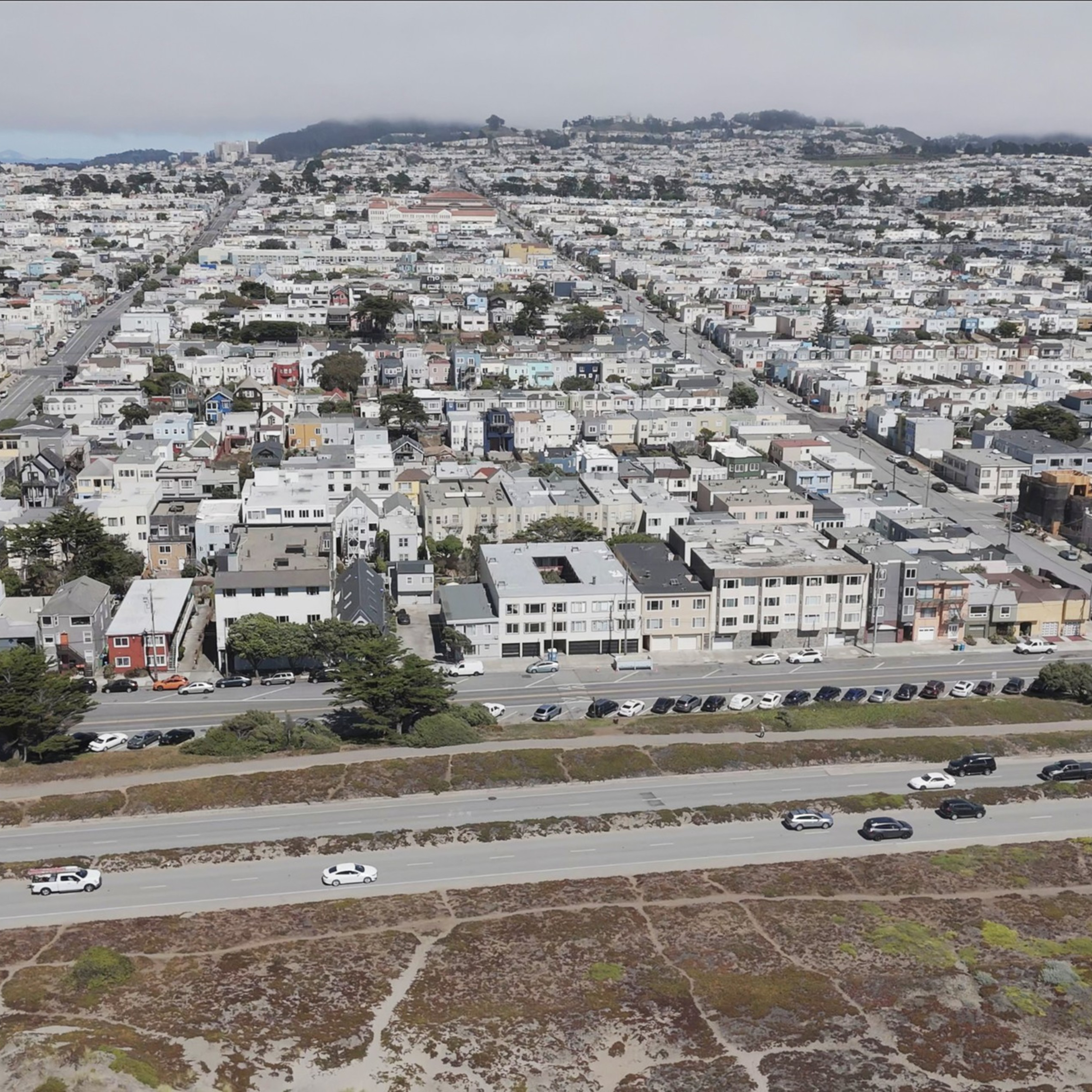 The image shows a densely packed suburban area with rows of houses, intersecting streets, and a highway with cars in the foreground. A hilly landscape is in the background.