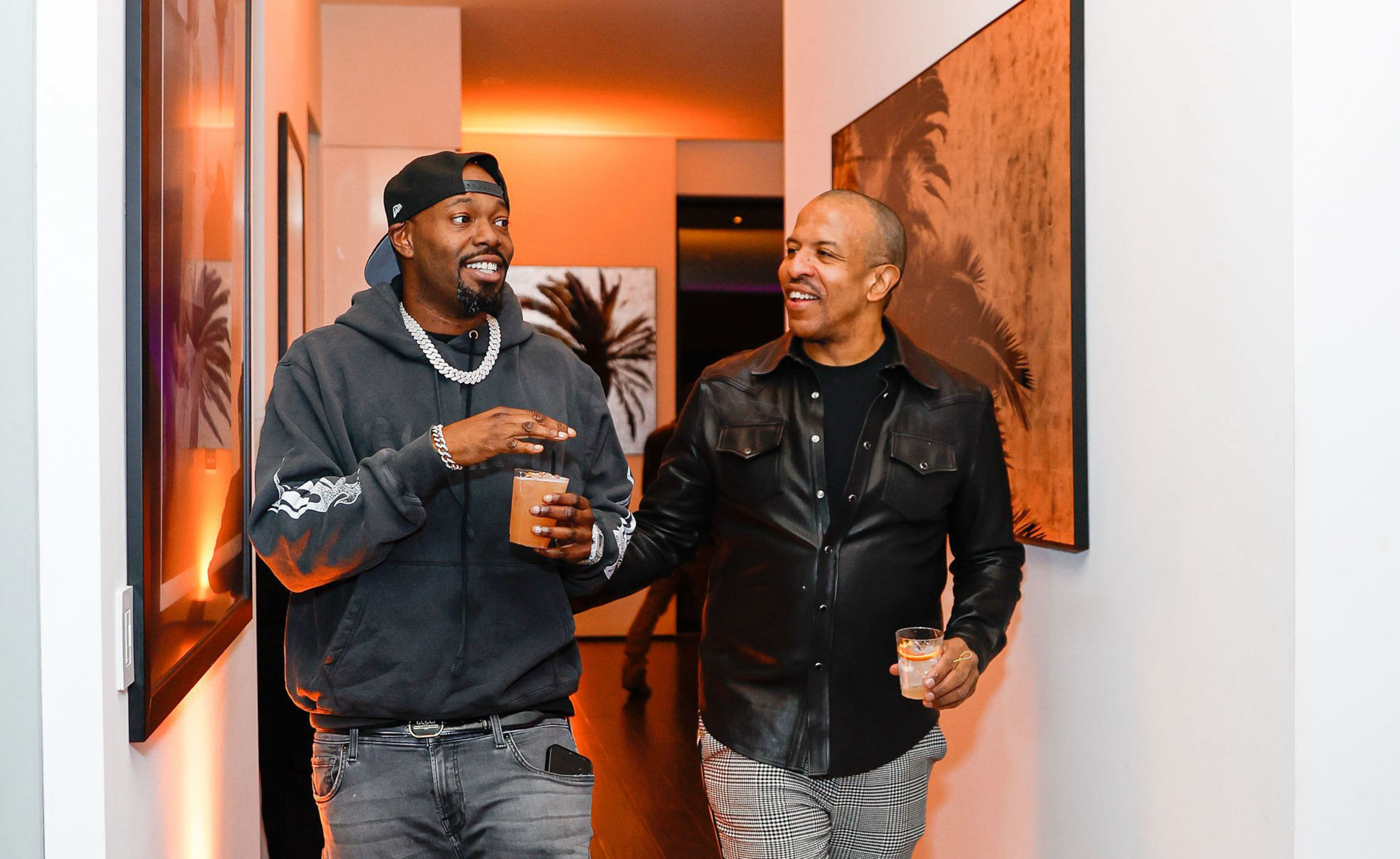 Two men walk down a hallway, smiling and holding drinks, with one wearing a black cap and hoodie and the other in a black leather jacket.