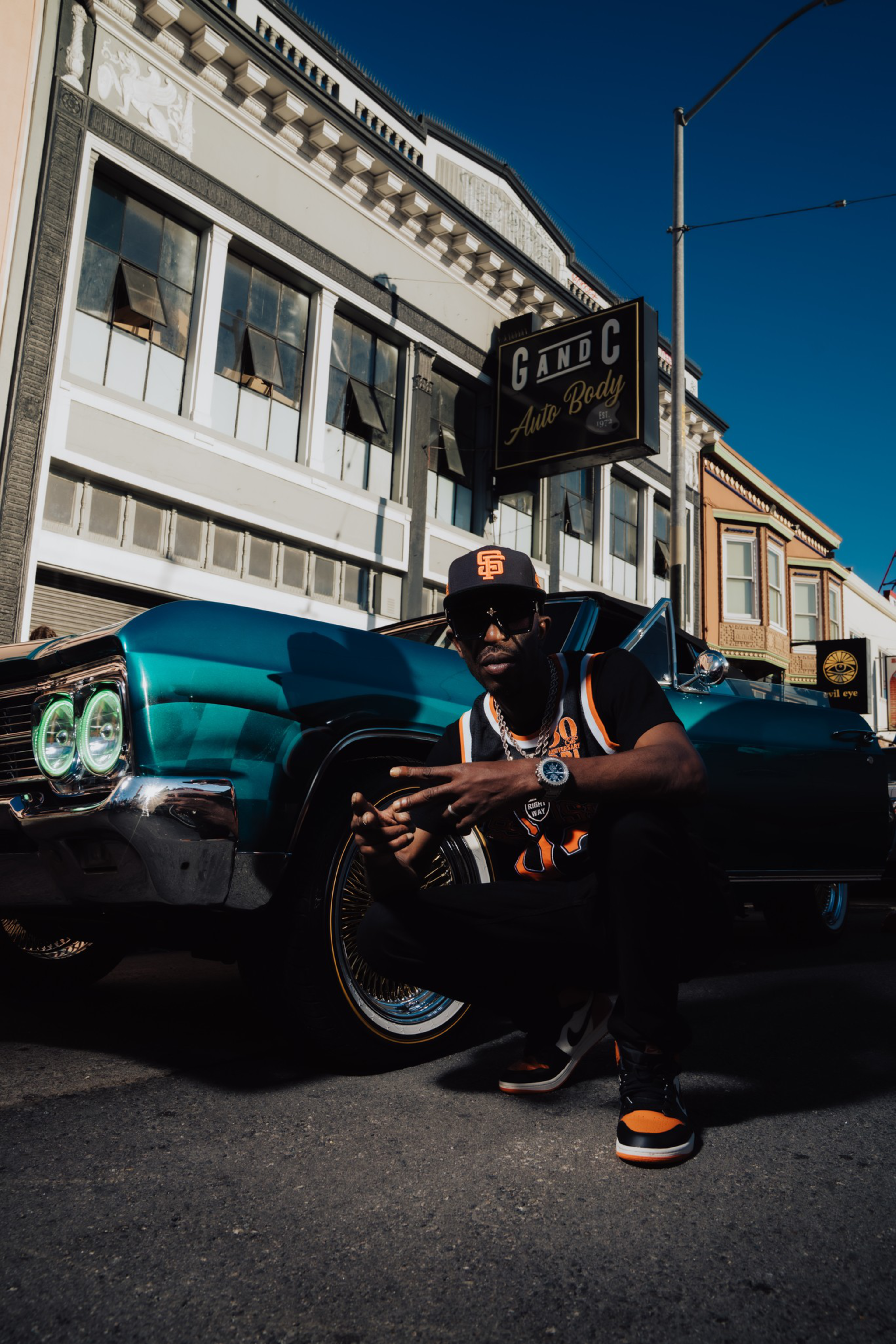 A man wearing a black and orange sports jersey and cap crouches beside a teal classic car with glowing headlights in front of a vintage auto body shop.