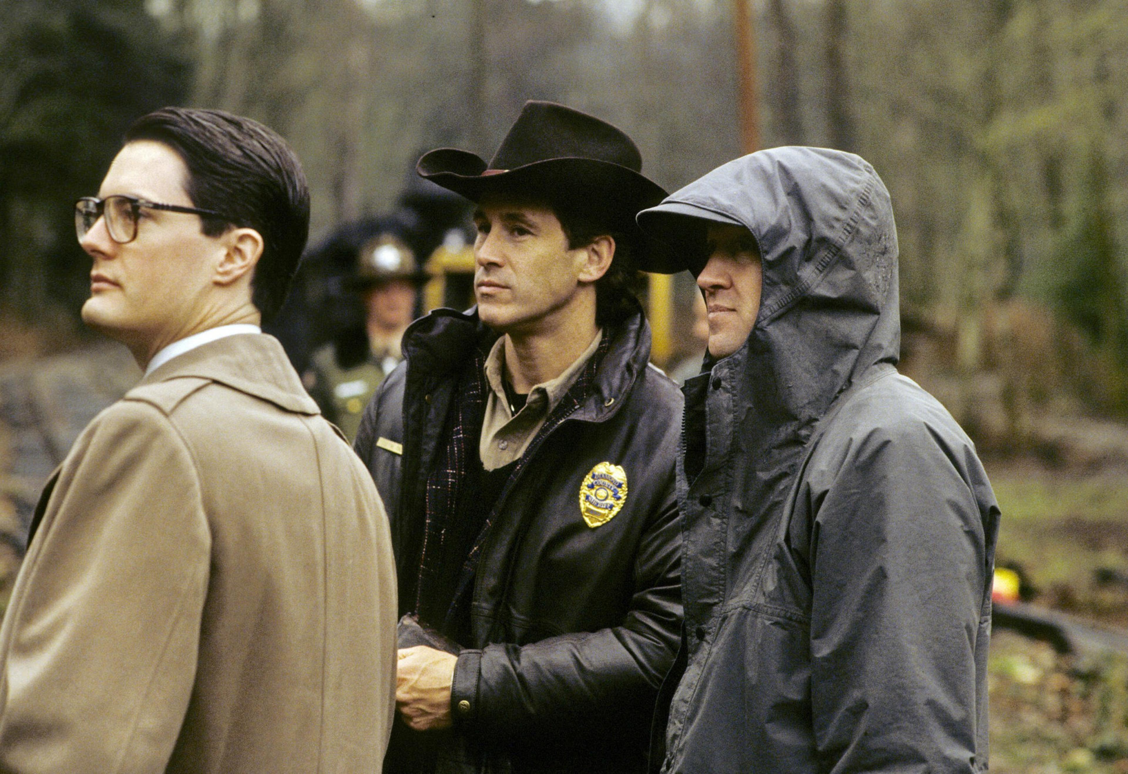 Three men stand side by side outdoors; one wears glasses and a tan coat, another a sheriff's badge and cowboy hat, and the third a hooded rain jacket.