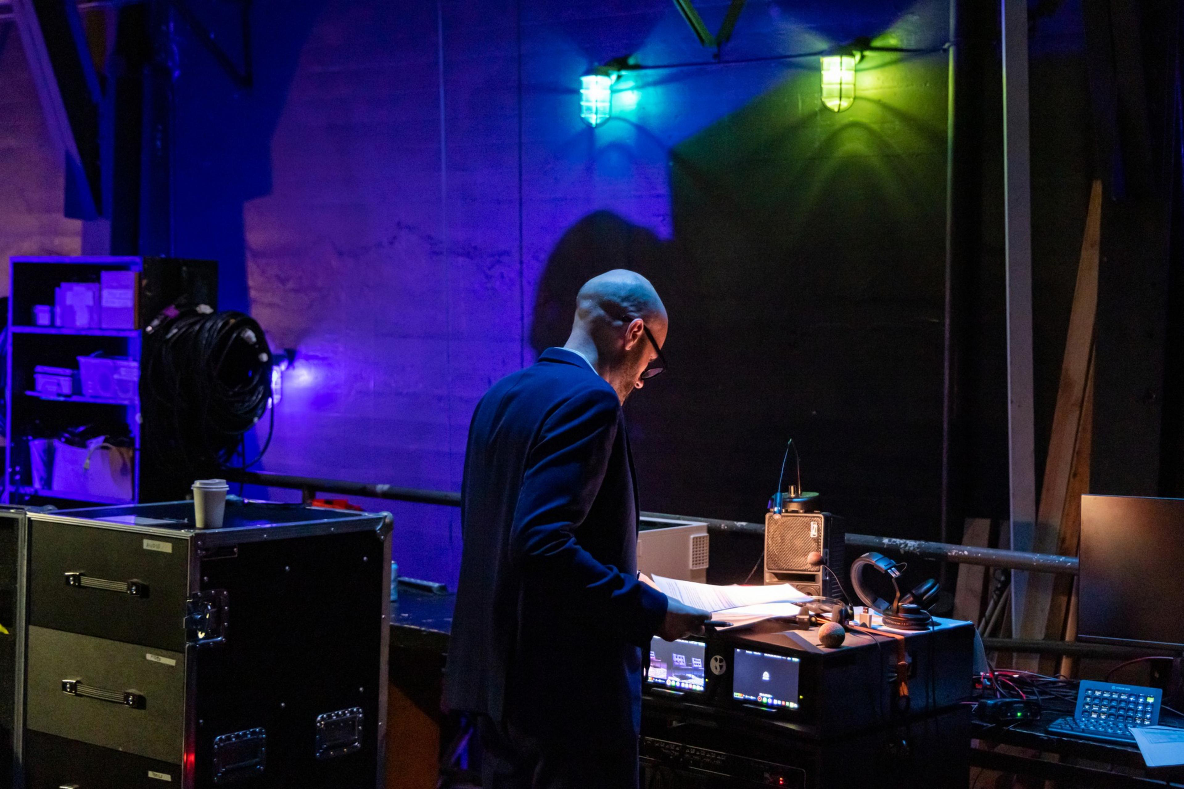 A person in a suit stands backstage in a dimly lit area, reviewing papers over sound equipment, with colorful lights illuminating the scene.