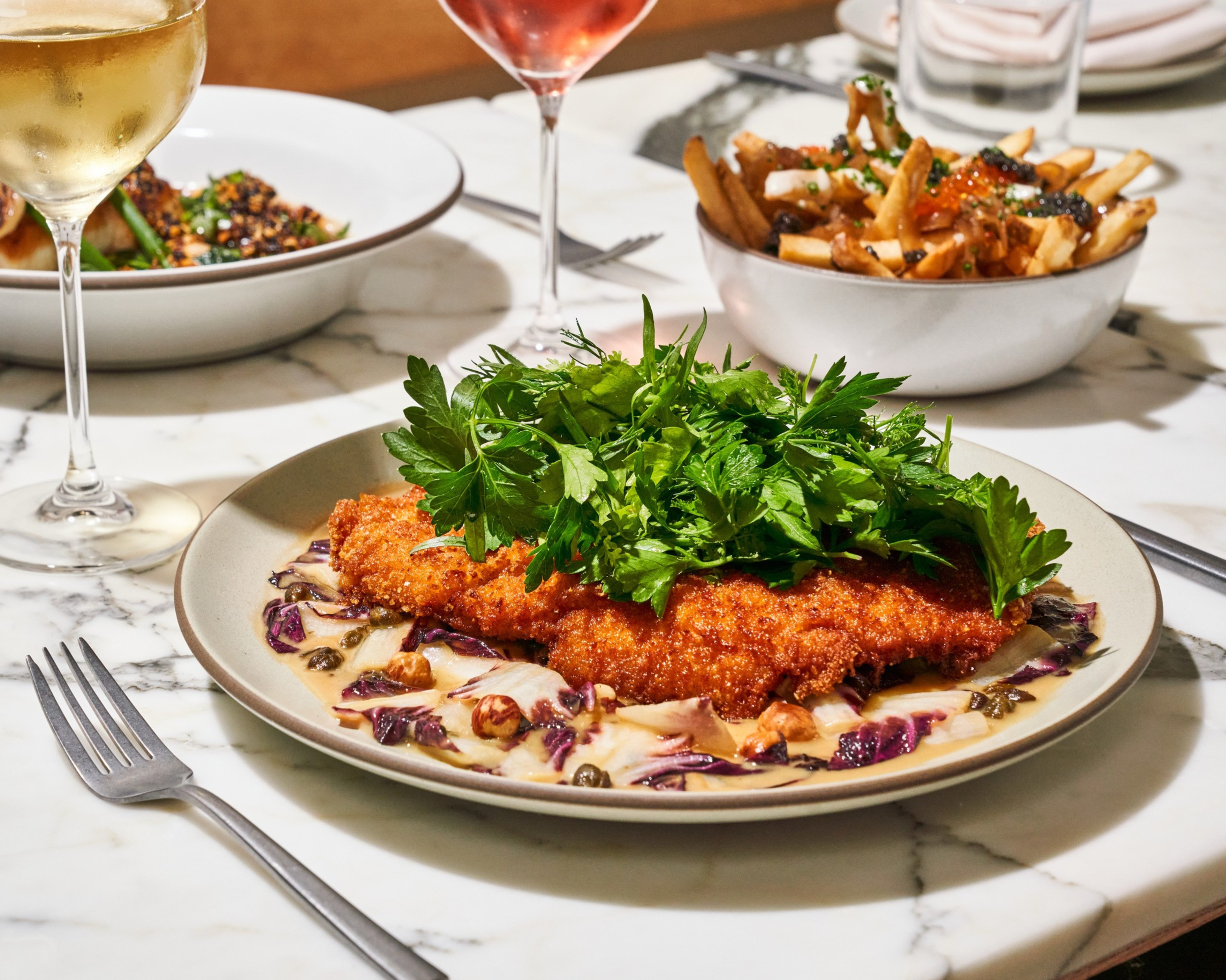A plate of crispy fried food garnished with leafy greens sits on a marble table, alongside dishes of fries and a hearty stew. Two glasses of white and rosé wine are nearby.