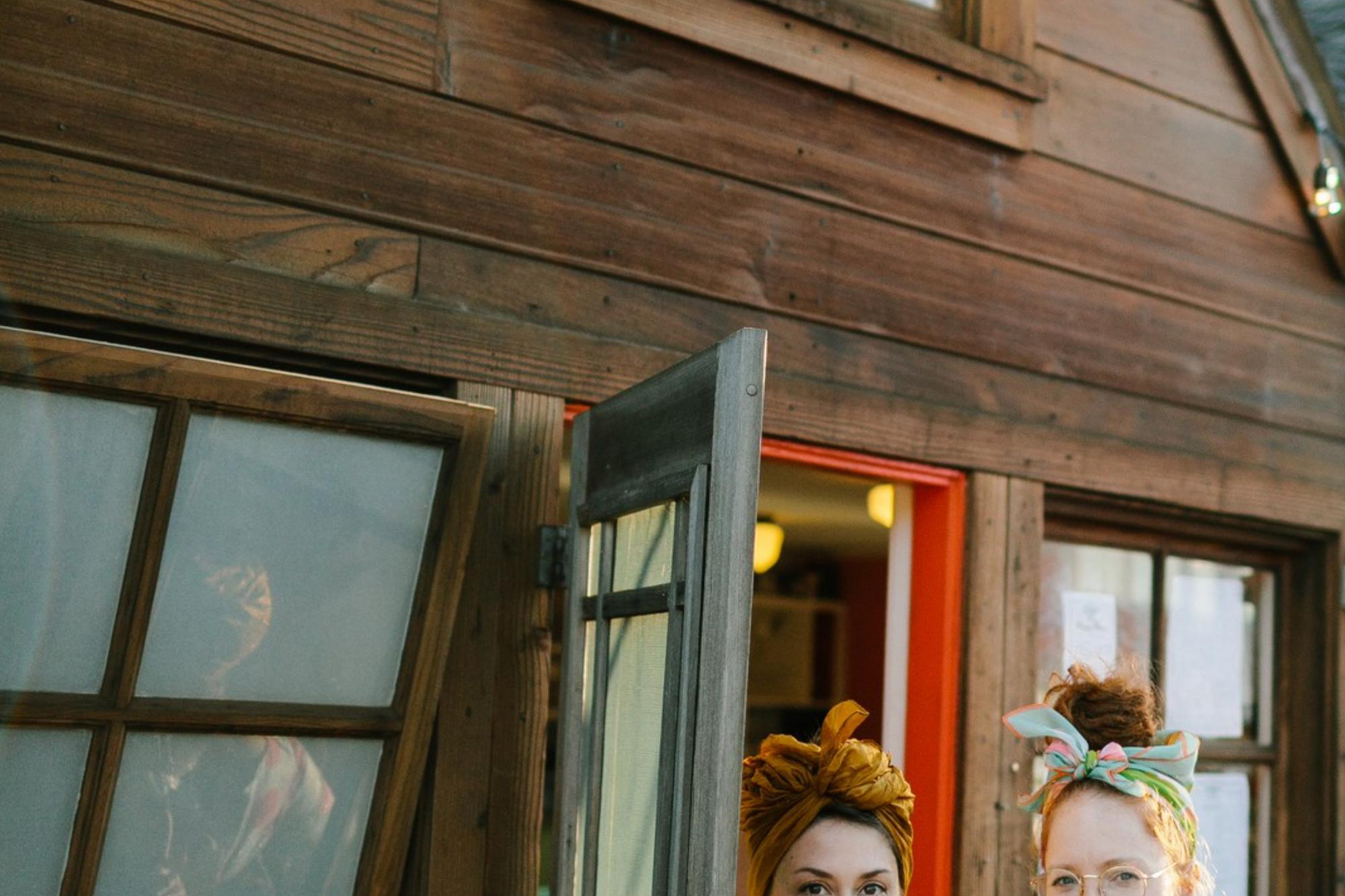 Two people stand smiling outside a rustic cafe, holding drinks. The sign reads "Fog Eater Cafe". They're dressed vibrantly, with string lights above.