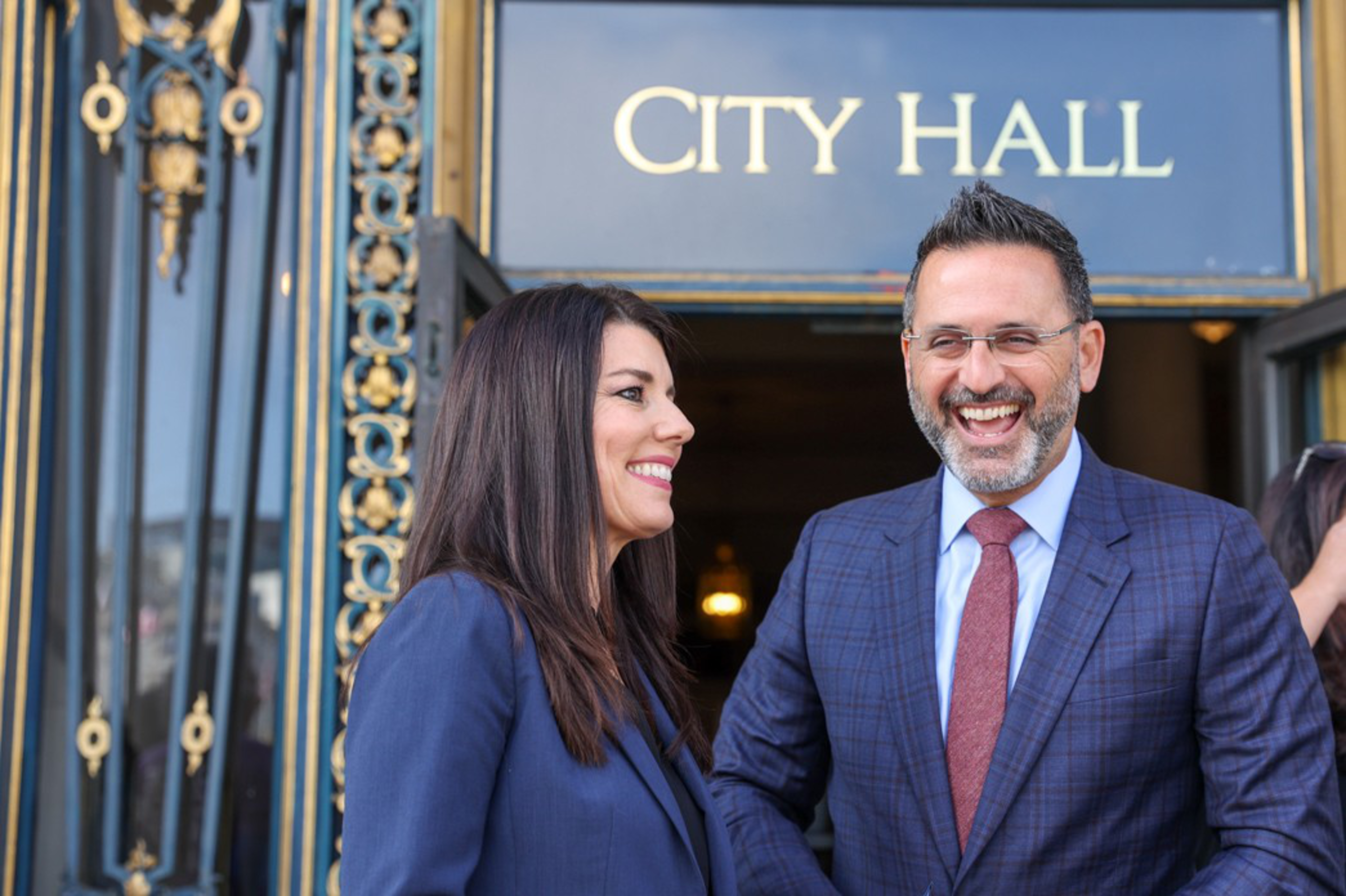 A man and woman in business attire are smiling and talking outside a City Hall entrance with decorative black and gold details.
