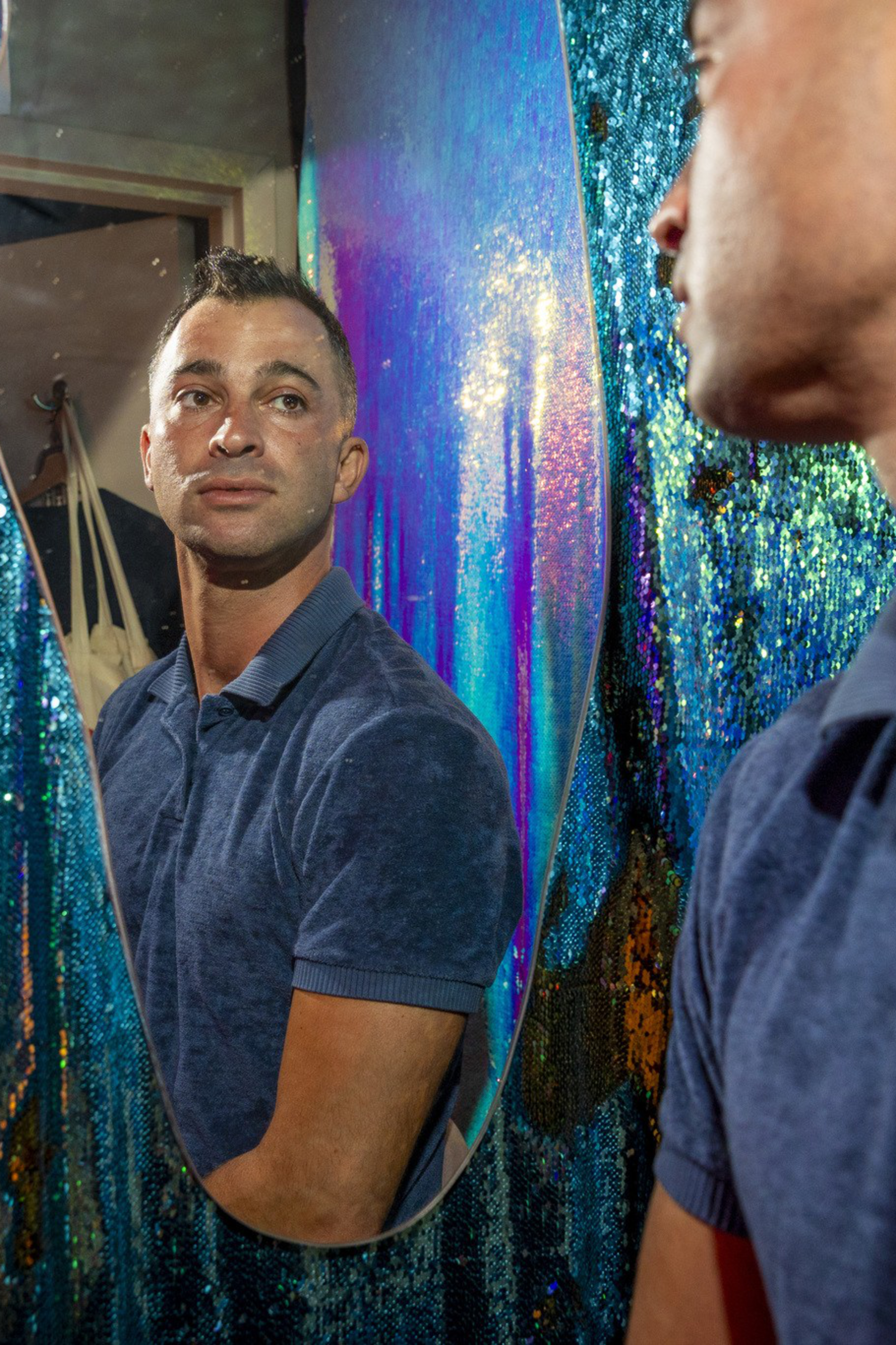 A man in a blue polo shirt looks at his reflection in a narrow, irregularly shaped mirror against a glittery, sequined blue and orange backdrop.