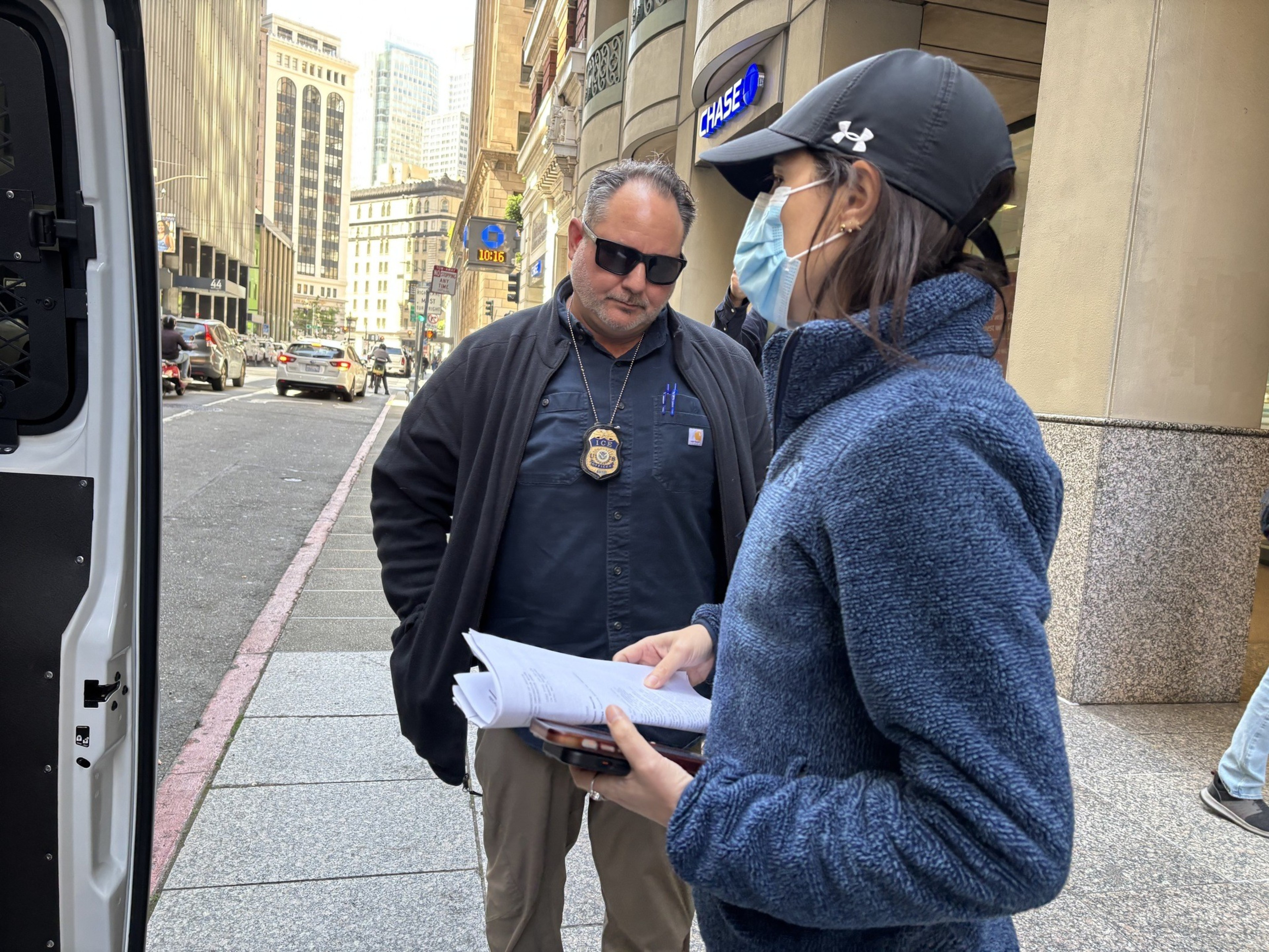 A man in dark clothing with a badge is talking to a woman in a blue jacket and cap, holding papers. They're on a city street near a Chase bank.