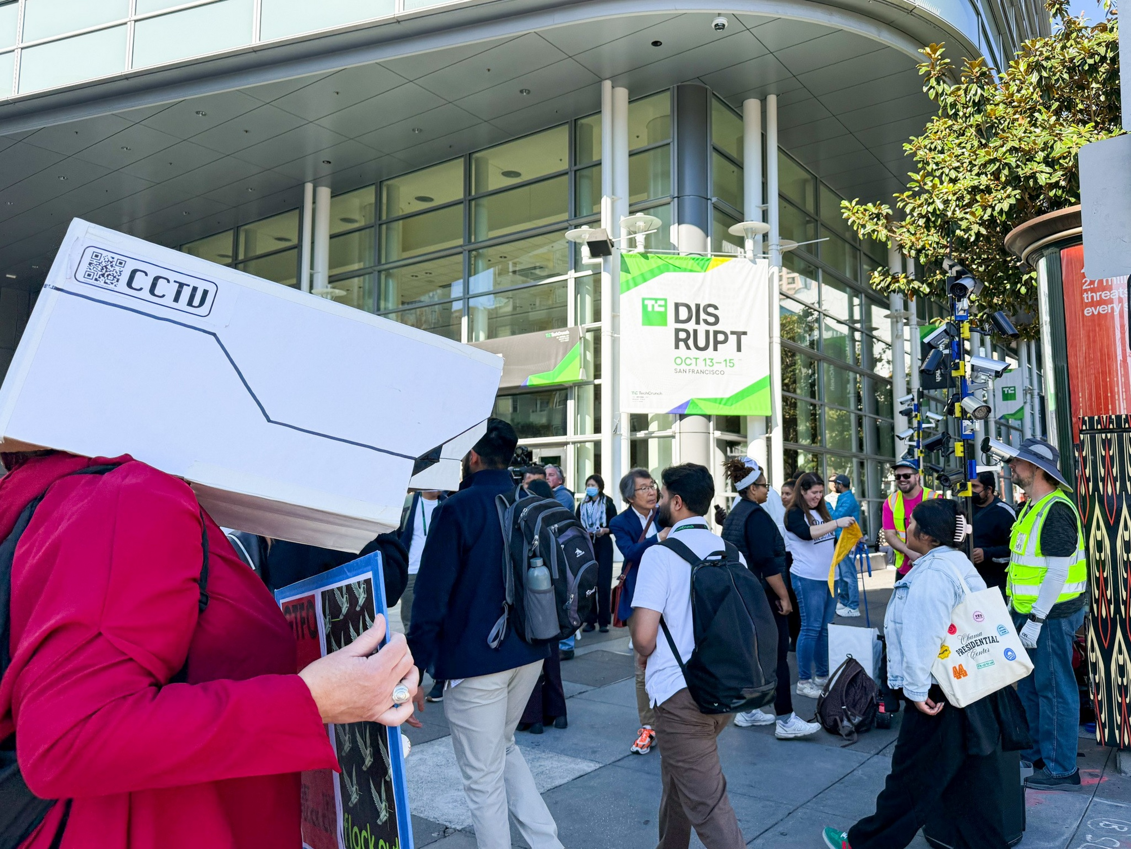 A person wearing a large mock CCTV camera head stands among a crowd outside a building hosting the TechCrunch Disrupt event in San Francisco.