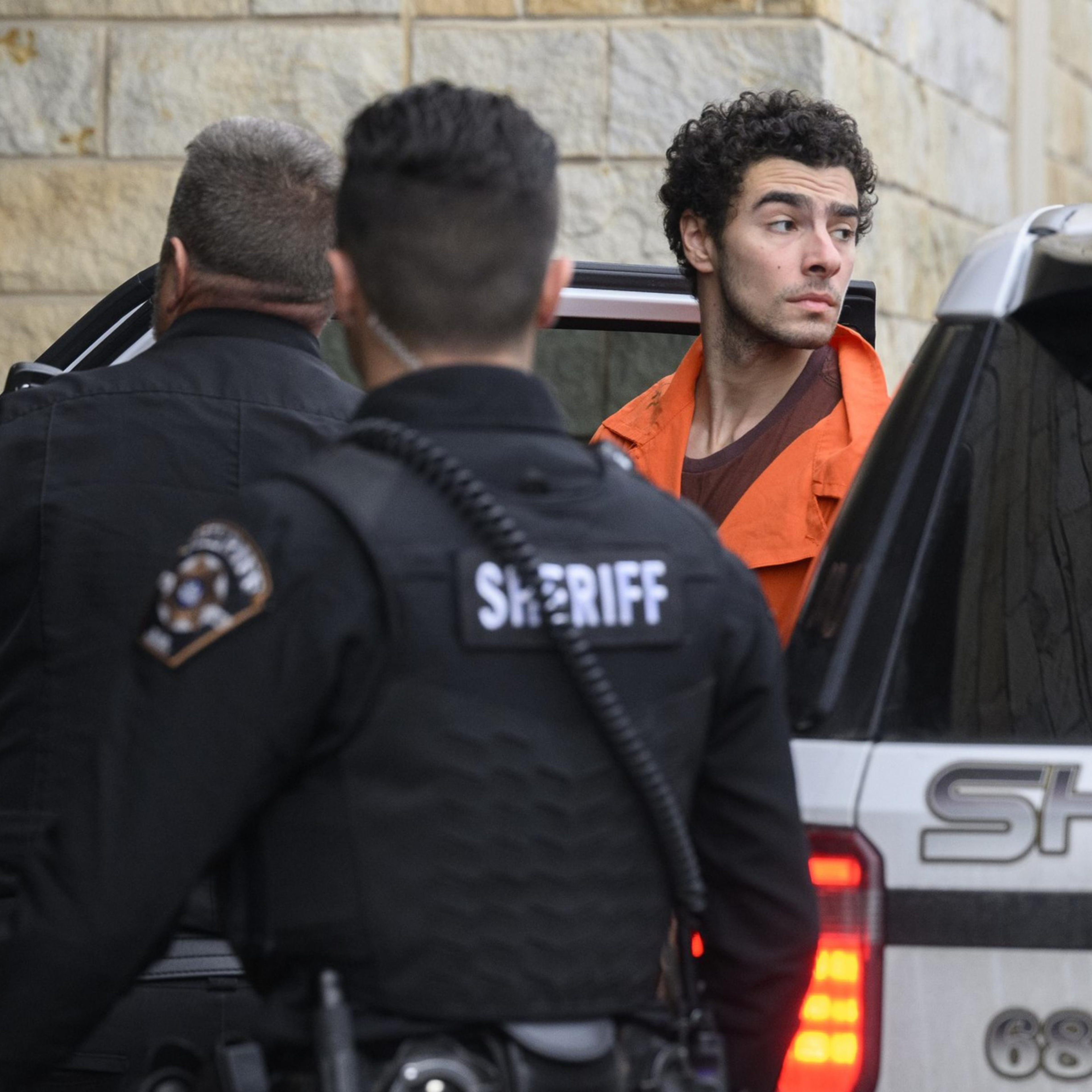 A man in an orange jumpsuit exits a sheriff's vehicle, accompanied by two officers in black uniforms. They are outside a stone building.