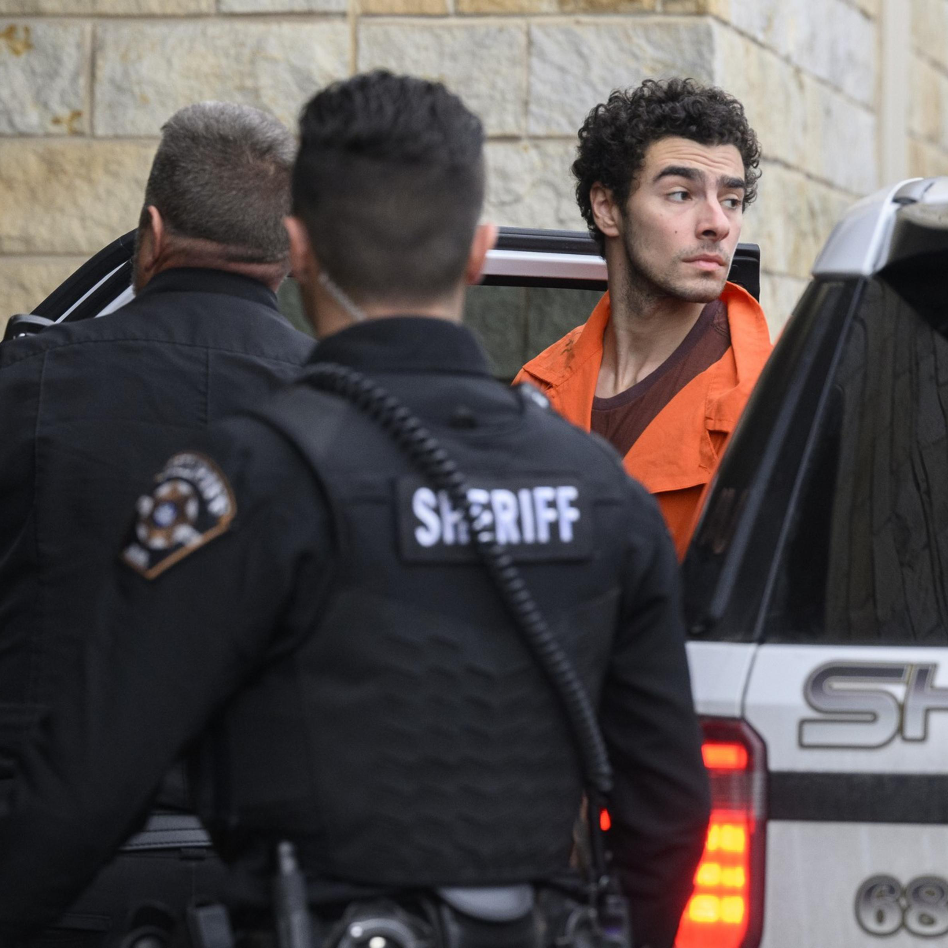 A man in an orange jumpsuit exits a sheriff's vehicle, accompanied by two officers in black uniforms. They are outside a stone building.