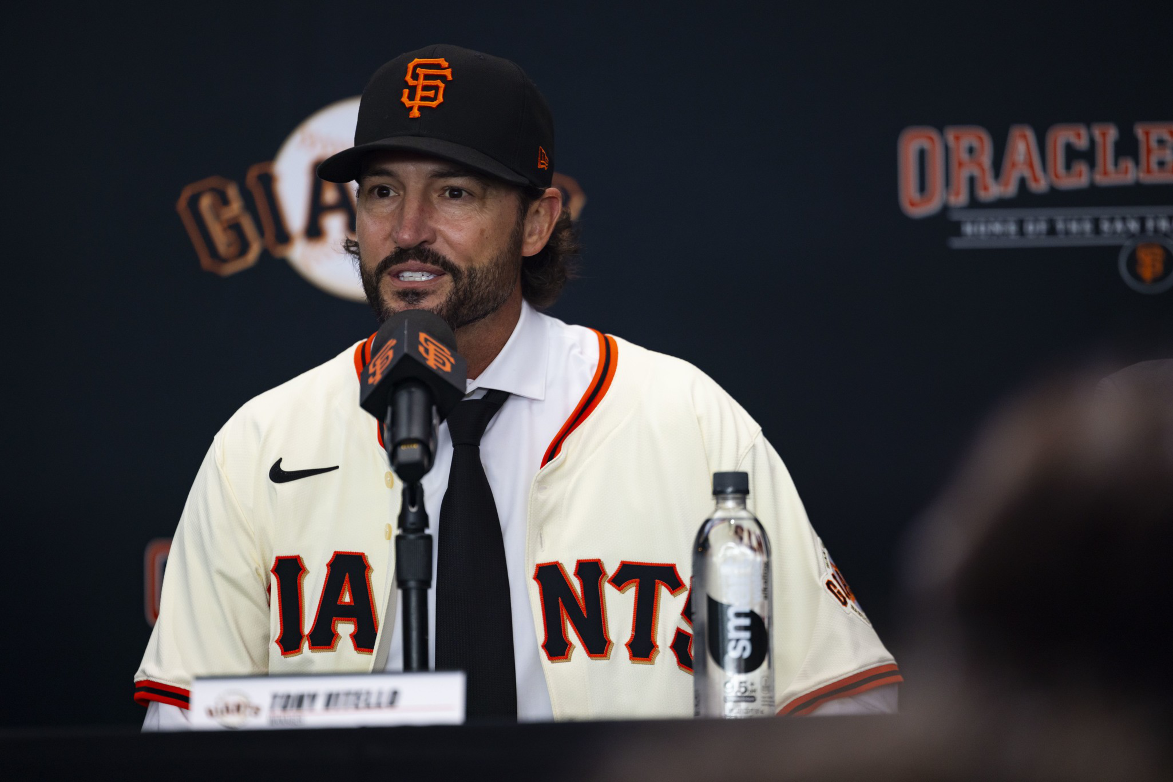 A man in a San Francisco Giants baseball cap and jersey speaks into a microphone at a press conference with a water bottle nearby.